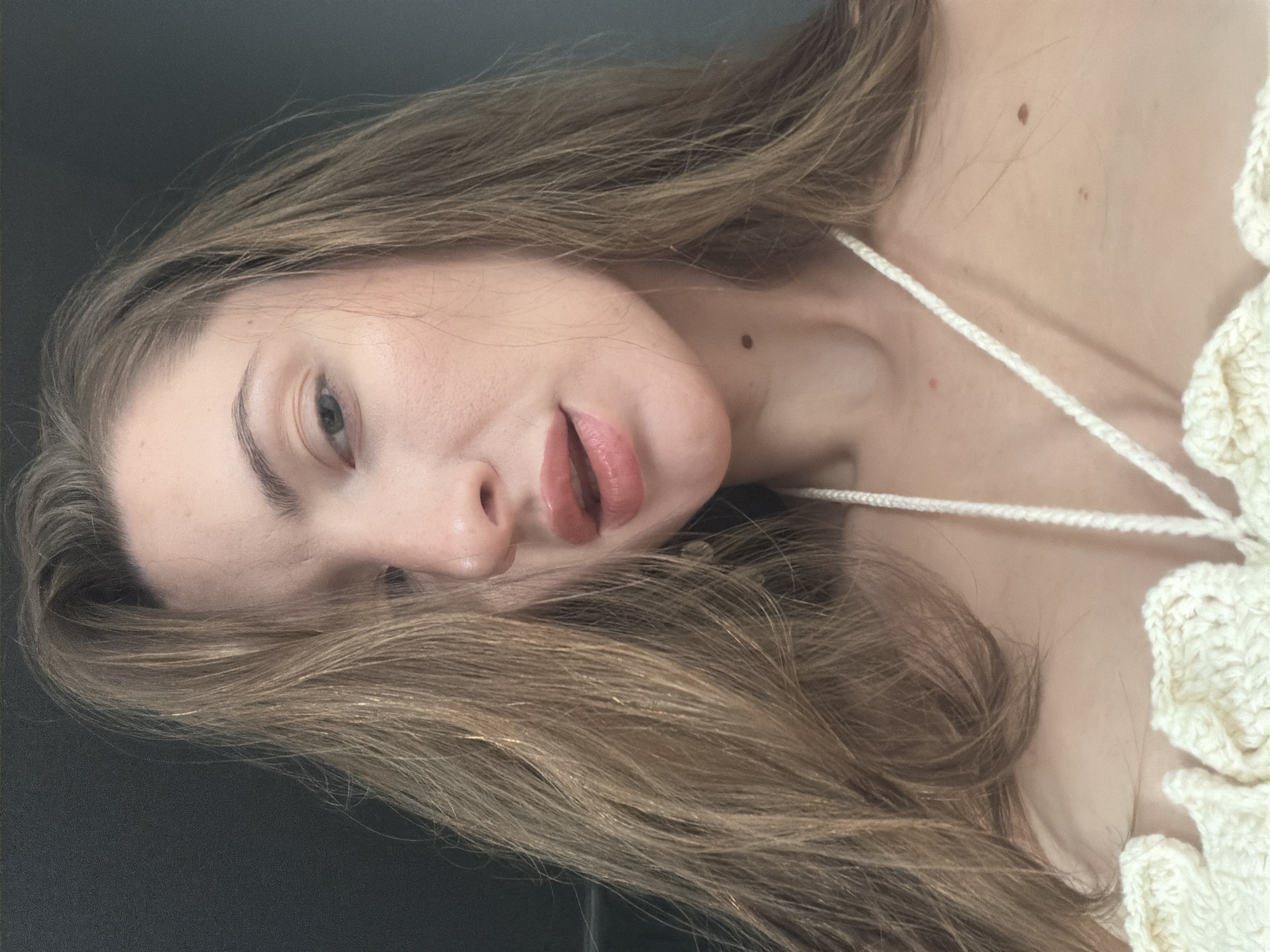 The image is a close-up, low-angle selfie of a woman with long, light brown hair. Her head is tilted, and her gaze is directed slightly upwards and to the left. She is wearing a crocheted off-white top. Her lips are slightly parted and appear to have a light lip gloss. A dark mole is visible on her neck. The background is a dark, textured surface. The lighting appears to be from the front. There is no indication of the location being Sharm El Sheikh, Egypt.