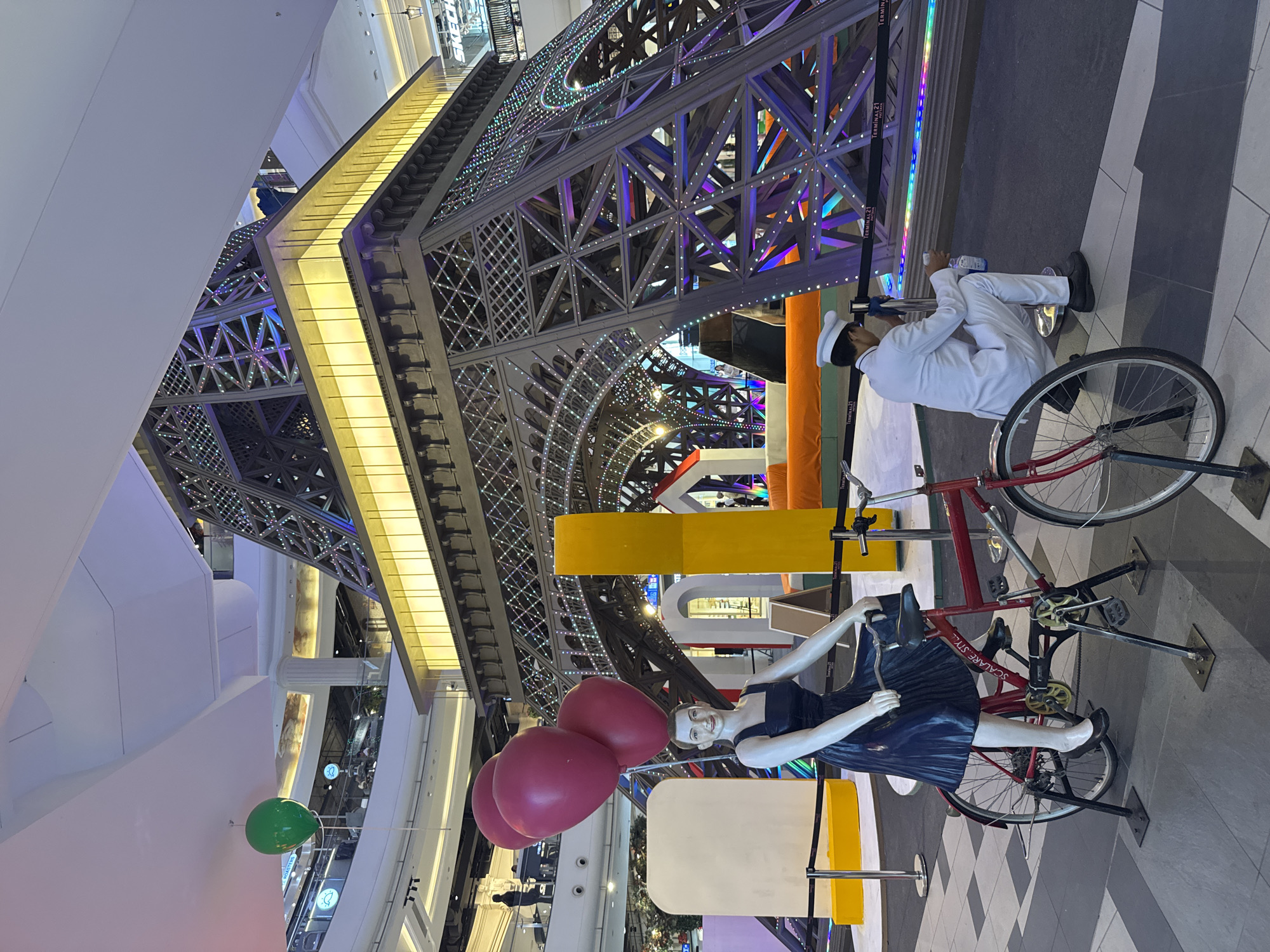 In Pattaya City, Thailand, this image captures a scene inside a shopping mall, likely Terminal 21, as indicated by the visible signage. A miniature replica of the Eiffel Tower, adorned with colorful LED lights, serves as the central attraction. In the foreground, a life-size mannequin depicts a woman riding a bicycle, with three pink balloons floating above her head. The mannequin is dressed in a dark blue dress and black flats. To the right of the mannequin, a man in a white uniform, including a white cap, is crouched down, holding a bottle. The flooring is tiled with a checkered pattern of dark grey and light grey squares. The overall environment suggests a festive or promotional display within a commercial space.