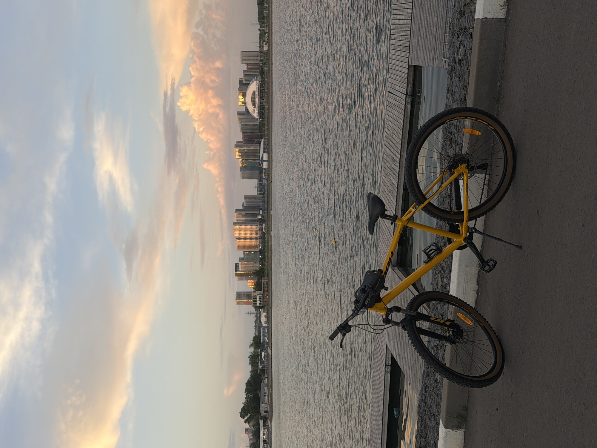 The image shows a yellow mountain bike parked on a paved surface next to a wooden dock. Beyond the dock is a body of water reflecting sunlight and the cityscape of Rostov-on-Don, Russia. The city skyline is visible in the distance and consists of several high-rise buildings. The sky is light with scattered clouds that are tinged with orange and pink hues. The bike is in the foreground, angled slightly towards the water.