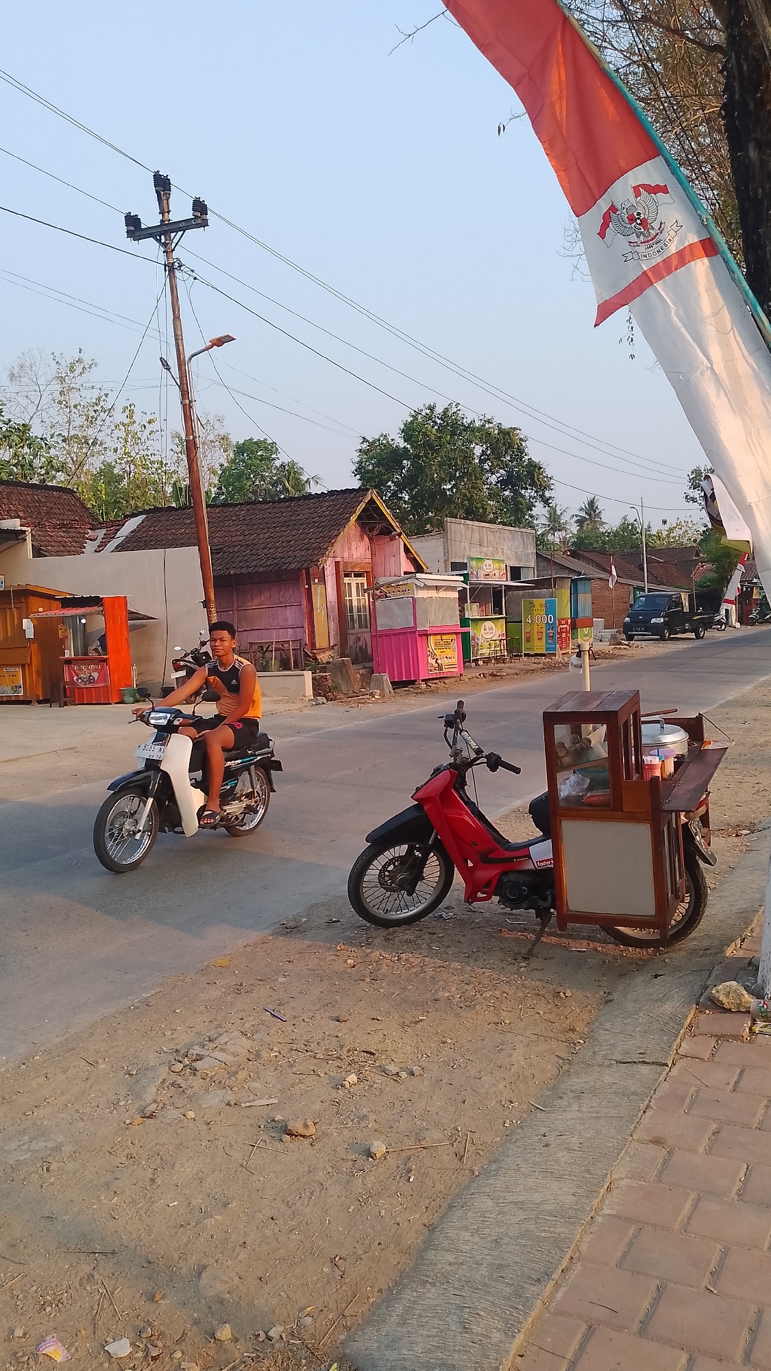 In the city of Karanglangu, Indonesia, on a dusty, sunlit street, a young man rides a black and white motorcycle. He is wearing an orange tank top and black shorts. In the foreground, a red motorcycle is parked, equipped with a mobile food cart made of wood. The cart has glass panels and appears to be set up for selling food, with a pot and various containers visible inside. In the background, small buildings line the street, some with colorful facades, suggesting a commercial or residential area. A utility pole with power lines stands to the left, and a large Indonesian flag, red and white, is partially visible in the upper right corner, adding a nationalistic element to the scene. The sky is clear and blue, indicating a warm, bright day.