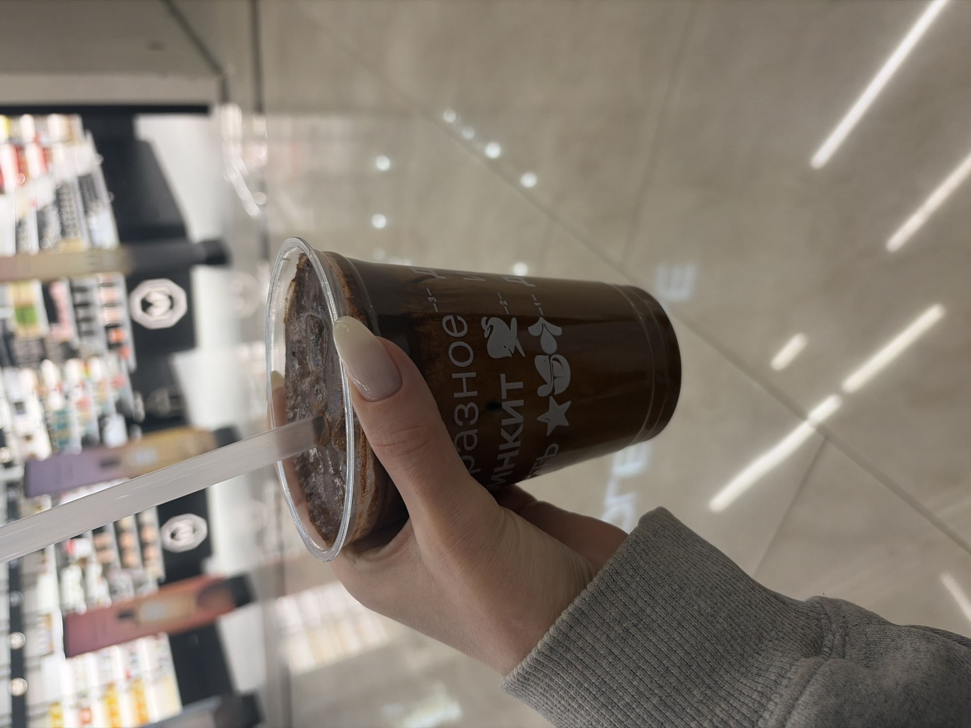 A close-up, first-person perspective photograph depicts a right hand holding a clear plastic cup containing a dark brown, viscous beverage, possibly a chocolate or coffee mixture with visible texture. A translucent white straw is inserted into the drink. The visible portion of the arm is clad in a light grey, ribbed long-sleeved garment. The fingernails are manicured with an almond shape and a light, neutral polish.
The cup features white Cyrillic text and stylized graphics. Clearly visible text includes "разное" (raznoye), meaning "various" or "different," and "МИНКИТ" (MINKIT), identifying the beverage brand. Below this, a star and a stylized creature (resembling a whale or fish) are printed, alongside measurement lines marked "-3-", "-2-", and "-1-".
The background is intentionally blurred, revealing a brightly lit indoor retail setting. Out-of-focus shelves stocked with various products, potentially cosmetics or personal care items, are visible, along with two tall, narrow display stands featuring graphic designs. The floor is composed of reflective, light-colored tiles, exhibiting prominent light reflections from overhead illumination.
Based on the "MINKIT" branding on the cup and the typical retail environment, the scene is located within a MINKIT establishment, likely situated inside a shopping mall or retail center, in Perm, Russia.