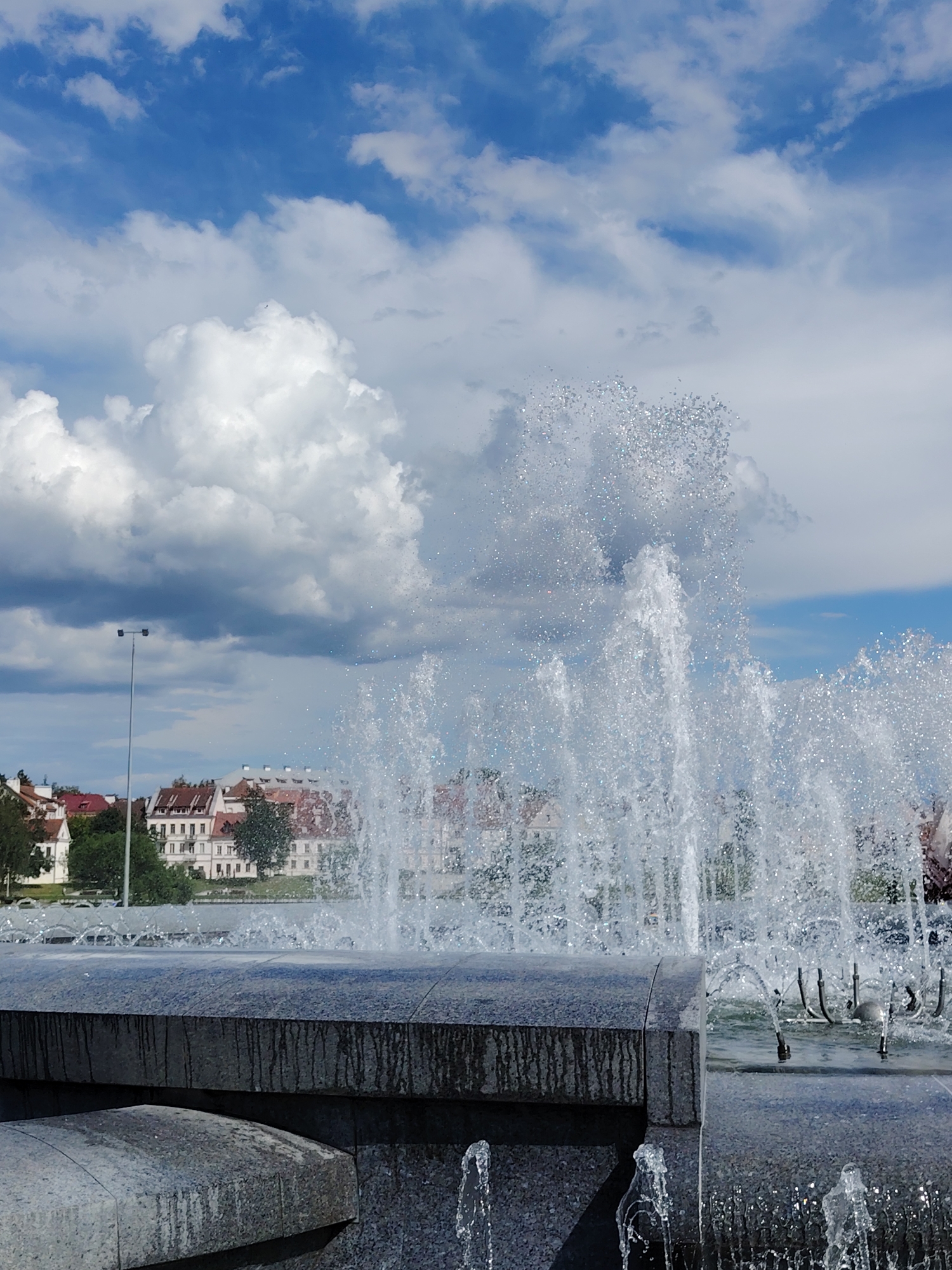This vertical outdoor shot captures a dynamic fountain against a backdrop of urban architecture and a bright, cloudy sky in Minsk, Belarus. The main subject is a large, multi-tiered fountain made of dark grey or black polished stone, occupying the foreground and midground. Numerous jets of water are actively spraying upwards, creating a dazzling display of sparkling droplets that catch the sunlight. The water cascades over the stone basin, with visible streaks of water running down its sides. Smaller jets are also visible at the base of the fountain and along its edges.
In the midground, behind the fountain, a row of buildings with distinctive red-tiled roofs and light-colored facades is visible. These multi-story structures are partially obscured by trees and the spray of the fountain. A tall, slender light pole stands to the left of the buildings.
The sky above is a vibrant blue, filled with a mix of fluffy white cumulus clouds and larger, greyer cloud formations. The lighting suggests it is daytime, likely a bright and sunny day with scattered clouds. No people or visible text are present in the image. The scene conveys a sense of refreshing outdoor activity in an urban setting.