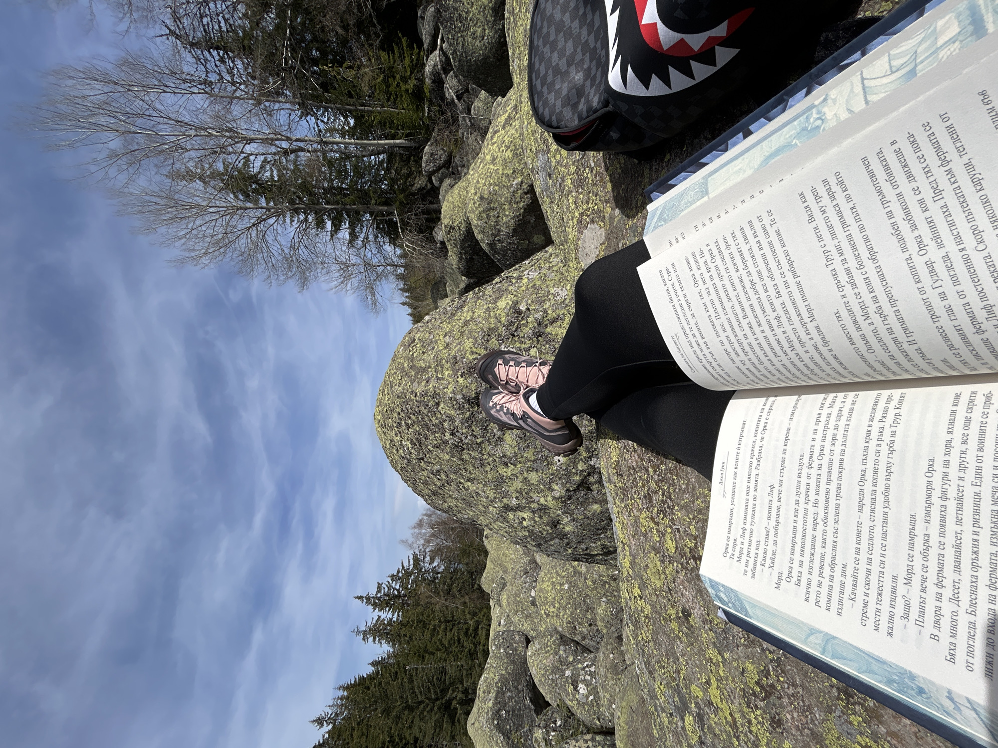 A person is sitting on lichen-covered rocks in a sunny, wooded area and reading a book. The person is wearing black leggings and pink and gray hiking boots. They have a book in their lap, open to a page of text written in Bulgarian. Next to the book is a black and gray bag with a red and white design. The rocks are surrounded by trees and other rocks. The sky is blue with some clouds.