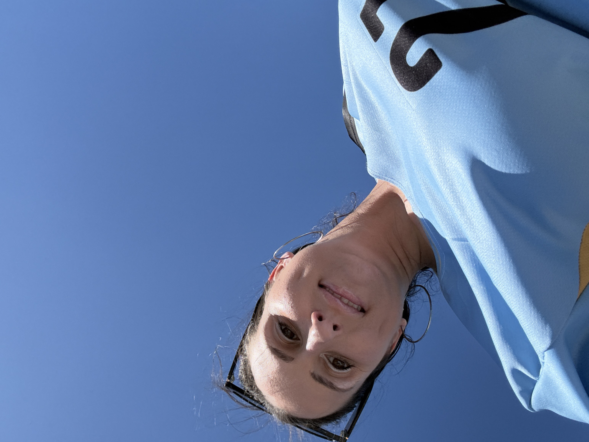 This is a low-angle, first-person selfie of a woman against a bright blue sky. The woman has dark hair, brown eyes, and is smiling. She is wearing a light blue t-shirt with black lettering on the front. She is also wearing large hoop earrings and sunglasses are pushed up on her head. The photo appears to have been taken outdoors on a clear, sunny day. No specific location in Miami Gardens is discernible from the image itself.