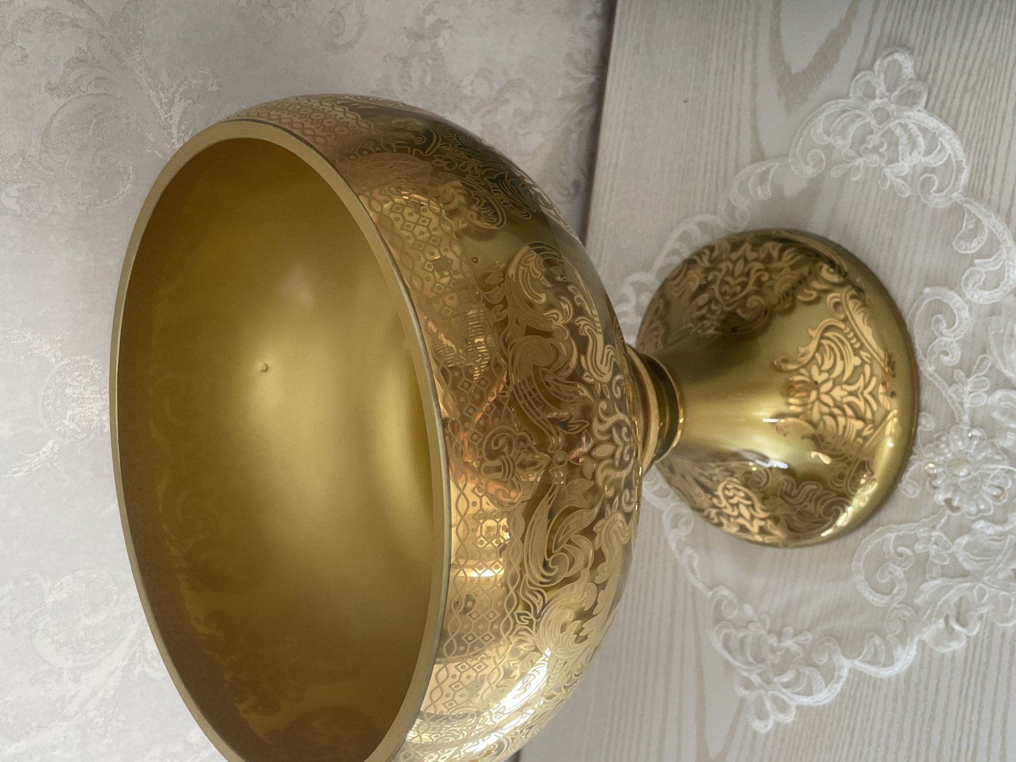 A golden decorative bowl with intricate patterns sits on a white lace doily, which rests on a light-colored wooden surface. The bowl has a wide, open top and a pedestal base. The background is a textured wallpaper with a light, floral pattern. The lighting suggests an indoor setting. There are no people visible in the image. The specific location in Tashkent, Uzbekistan, cannot be determined from the provided image.