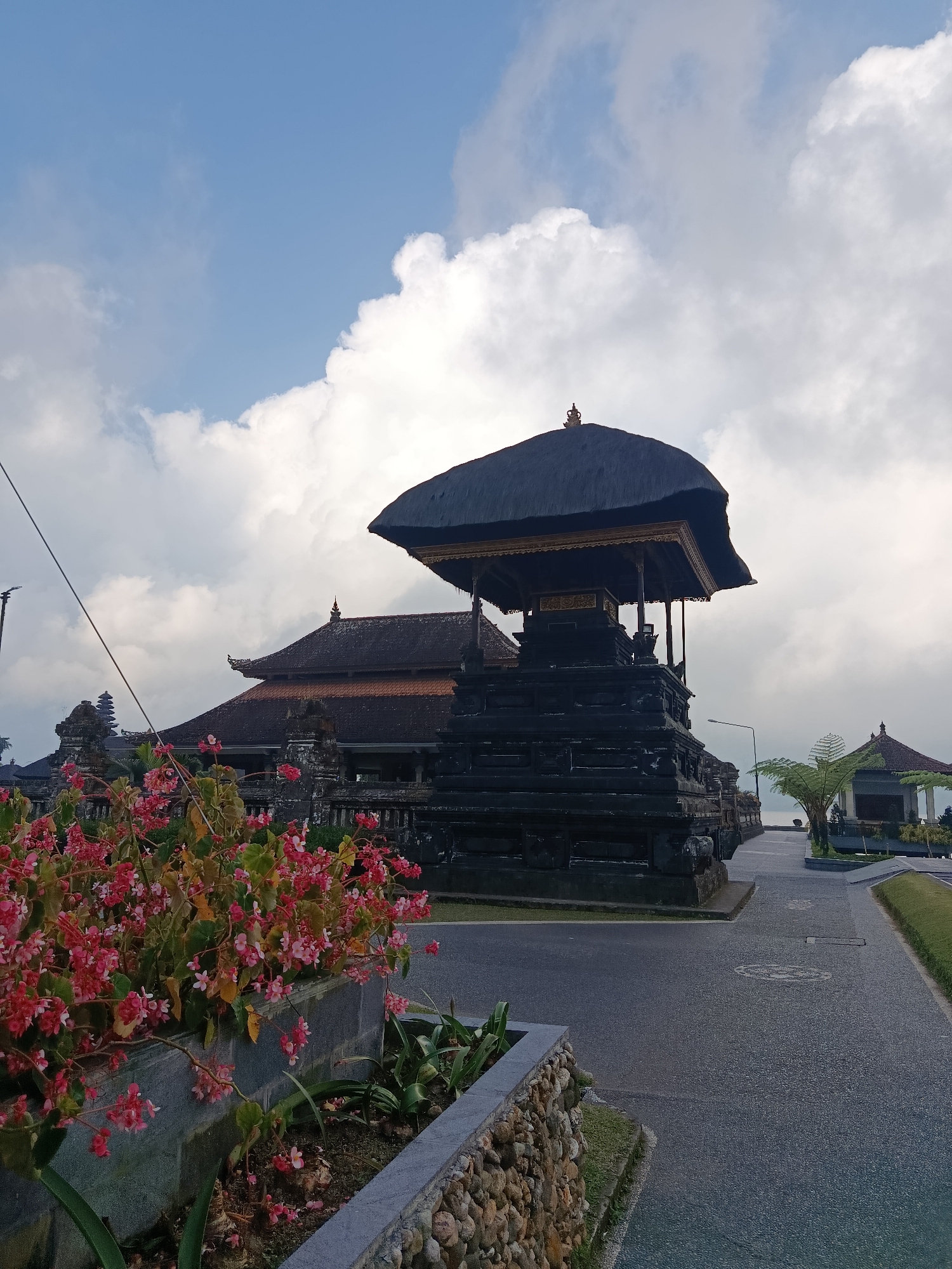 The image depicts the Pura Ulun Danu Batur temple complex in Baturiti, Indonesia. In the foreground, a planter box overflows with vibrant pink flowers and green foliage. Adjacent to the planter is a paved walkway leading into the temple grounds. The main subject is a dark, multi-tiered stone structure with a thatched roof, characteristic of Balinese architecture. Behind this structure, a larger building with a similarly dark, tiled roof is visible. The sky above is a mix of blue and white clouds. The overall scene suggests a serene, possibly early morning or late afternoon atmosphere, with the temple bathed in soft, diffused light.