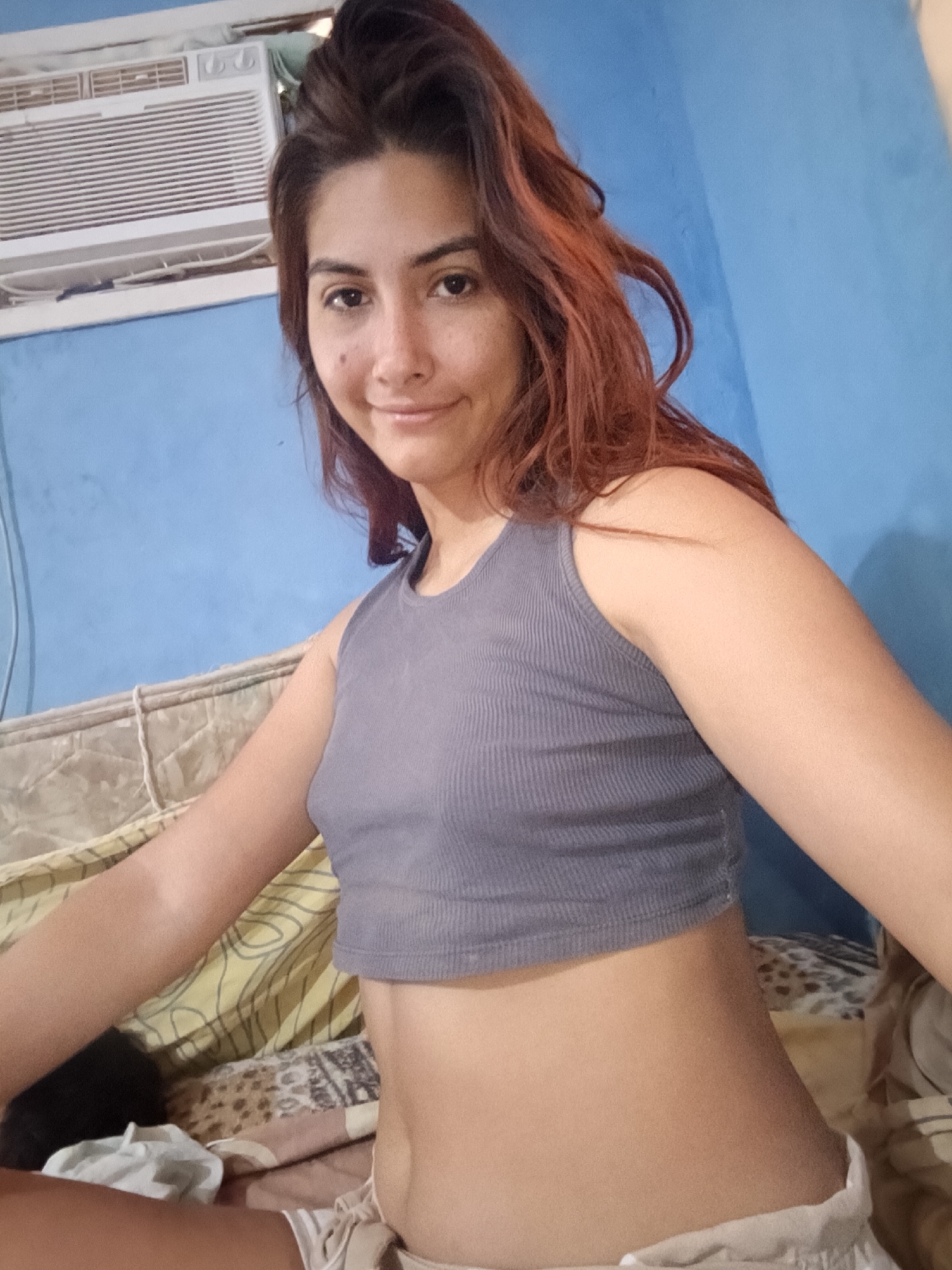 A medium shot depicts a woman with reddish-brown hair wearing a grey ribbed tank top. She is sitting on a bed in a room with blue walls. An air conditioner unit is visible at the top of the frame. The location appears to be in an indoor setting in an unknown city.