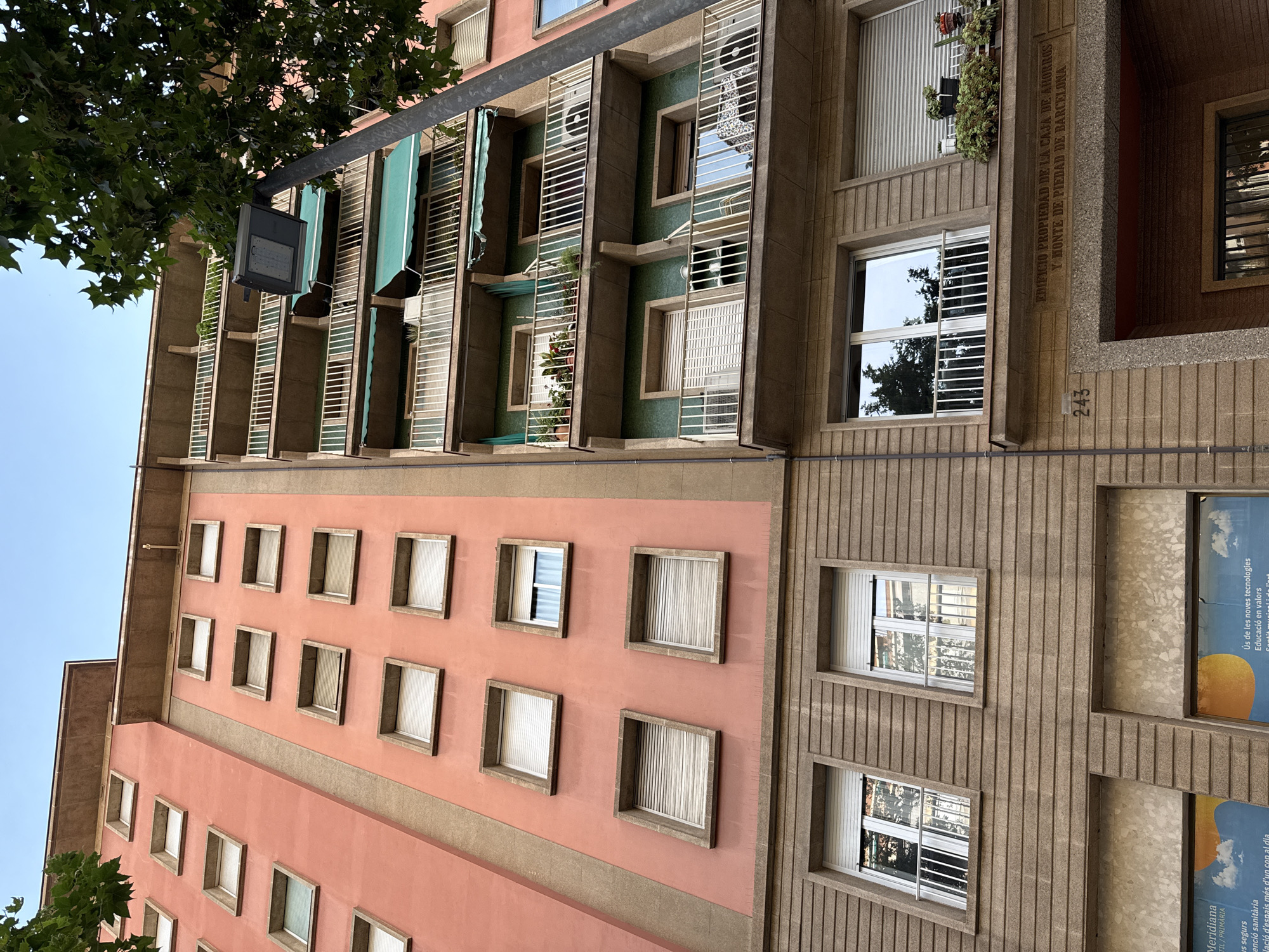 The image captures a low-angle view of two adjacent buildings in Barcelona, Spain. The left building is painted a vibrant coral pink with beige vertical trim. It features numerous square windows with light-colored blinds. The right building has a beige facade with balconies featuring white railings. These balconies are adorned with green retractable awnings and planters with greenery. The right building also has a distinct architectural element above the ground floor entrance that reads "EDIFICIO PROPIEDAD DE LA CAJA DE AHORROS Y MONTE DE PIEDAD DE BARCELONA" and the number "243" is visible on the facade. The sky above is a clear blue, and the top right corner of the image shows the leafy branches of a tree.