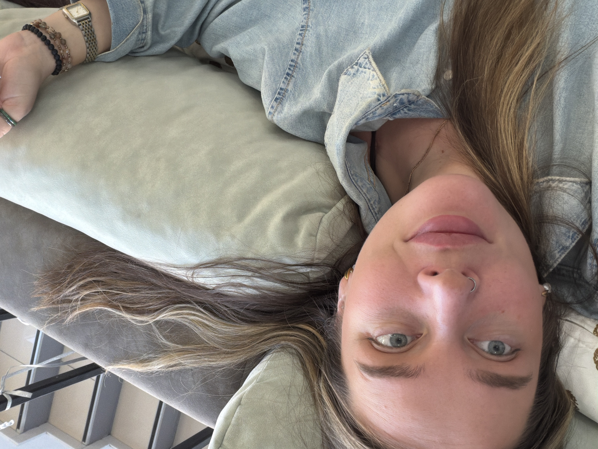 A woman with light skin, gray eyes, and light brown hair lies on a light green cushion with a gray trim. She has a nose ring and earrings. She is wearing a light blue denim shirt. Her arm is extended, adorned with a watch and bracelets. The floor and parts of the ceiling are visible in the background. The location is not discernable from the image.