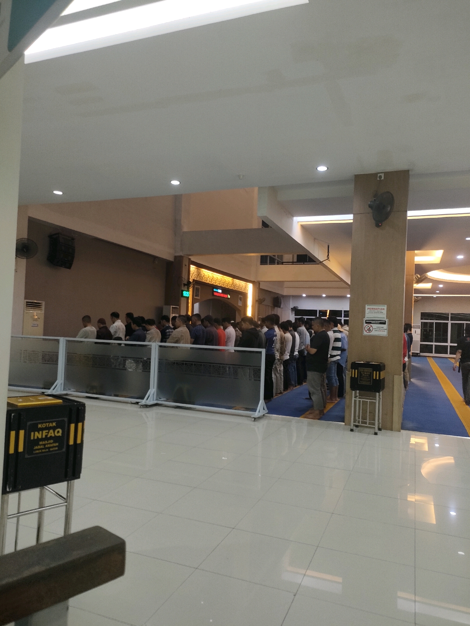 In a public space resembling a mosque or prayer hall in Batam, Indonesia, a group of men are lined up, facing a wall, engaged in prayer. The environment features a tiled floor, a patterned carpet, and overhead lighting. A donation box labeled "KOTAK INFAQ" is visible in the foreground, along with a metal stand and a sign in Indonesian reading "PERHATIAN" (Attention). The men are dressed in casual attire, and some appear to be younger individuals. The scene suggests a communal religious gathering.