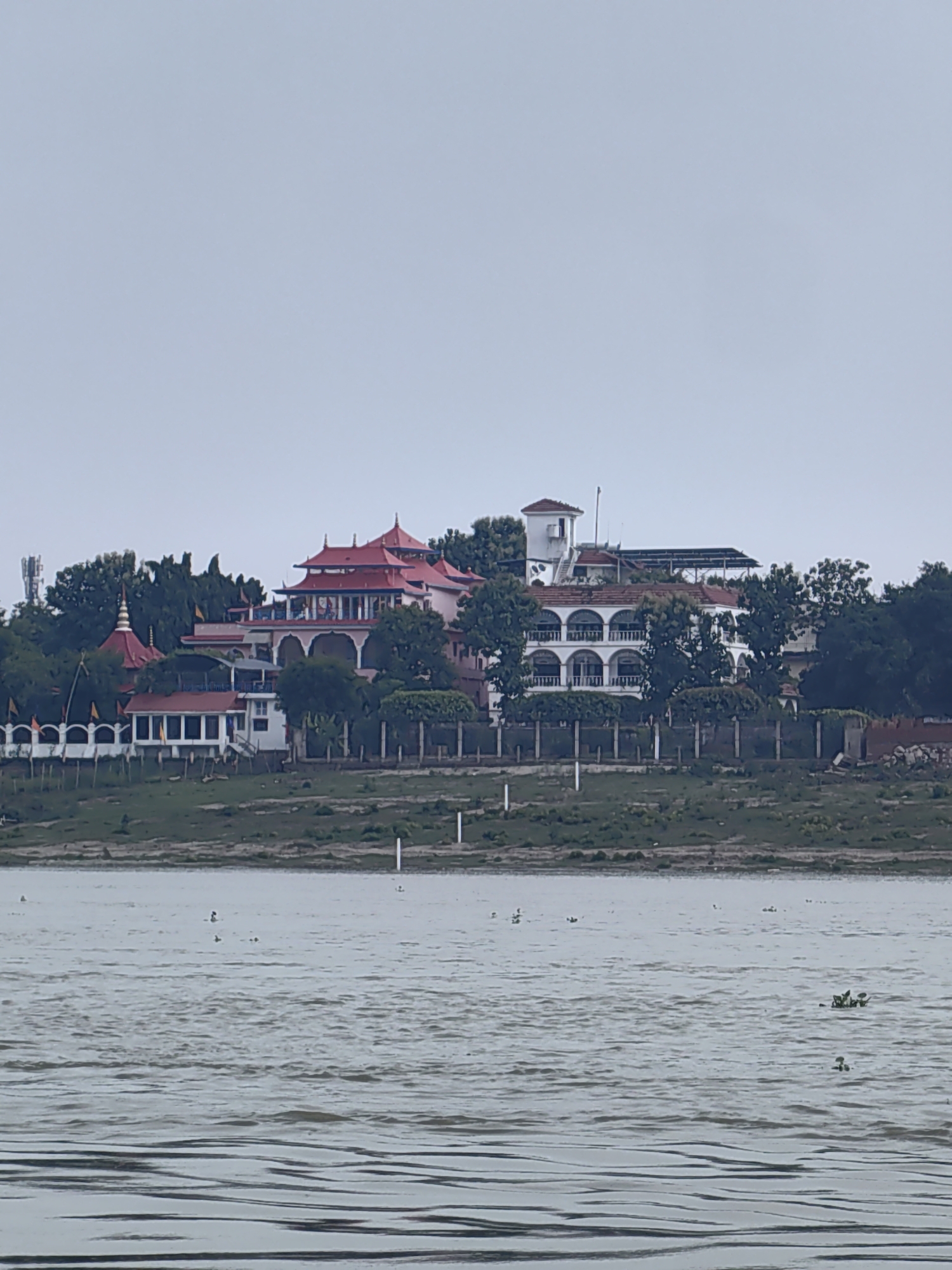 The image shows a waterfront view of buildings along the bank of a body of water, possibly a river. The structures appear to be a mix of traditional and modern architecture. On the left, there is a complex with multiple red-tiled roofs and white colonnades. A golden spire is visible above some of the buildings. Further to the right, a white building with arched windows and balconies is situated. Trees and greenery surround the buildings. The water in the foreground is rippling, and there are a few small plants floating on its surface, as well as what appear to be birds or ducks. The sky is overcast and a pale blue-gray. The overall impression is of a serene, possibly religious or historical site located in a humid, possibly tropical or subtropical environment. Given the context of Mughalsarai, India, this could be a temple complex or a historical building situated along the Ganges River.