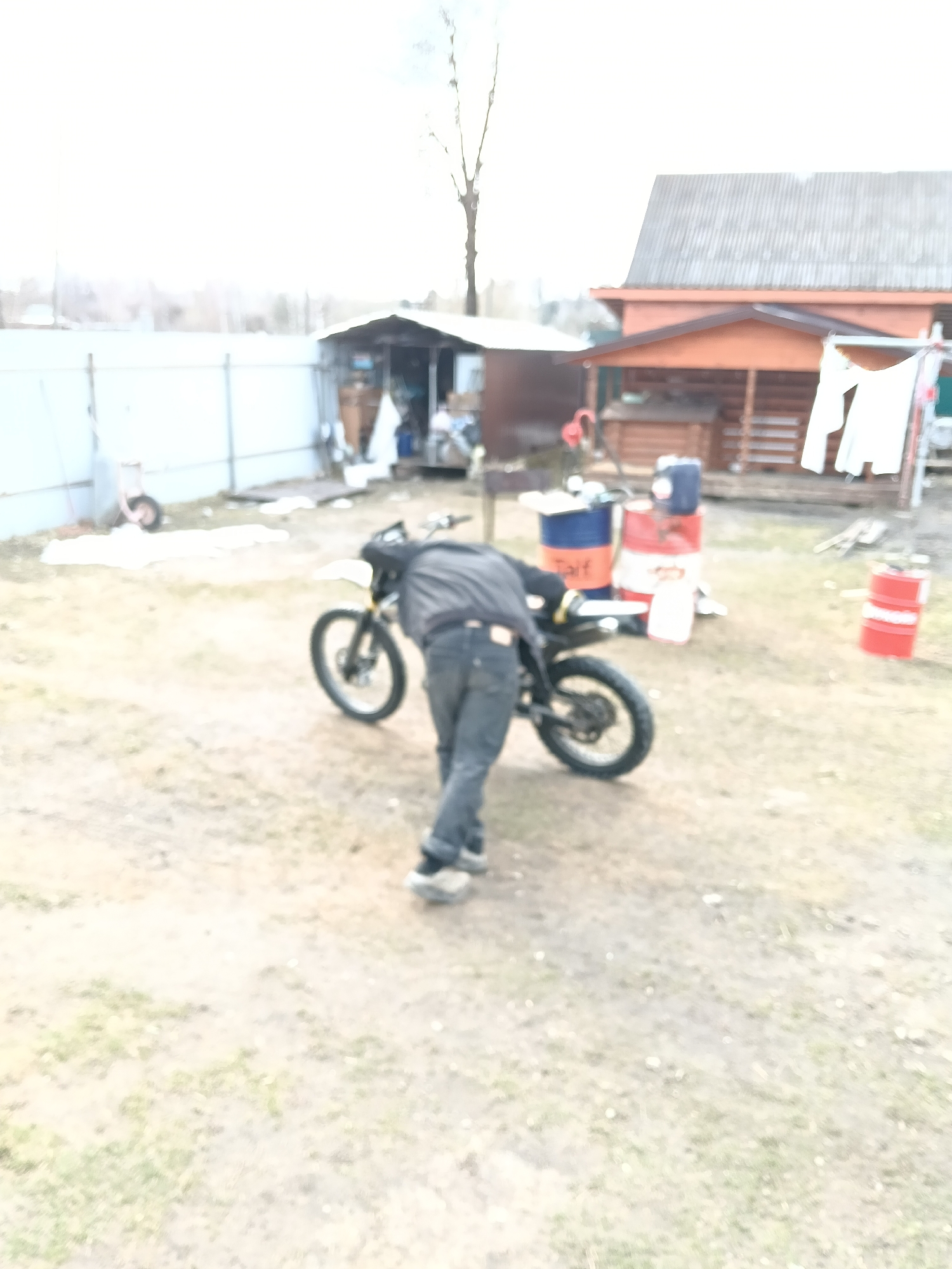 The image shows a blurry scene in Красный Маяк, Russia. An individual bends over a black motorcycle in a yard. Behind the motorcycle are several oil drums and containers. In the background, there's a wooden house, a shed, and a white fence. Clothing hangs on a line. The ground appears to be a mix of dirt and sparse grass. The sky is overcast.