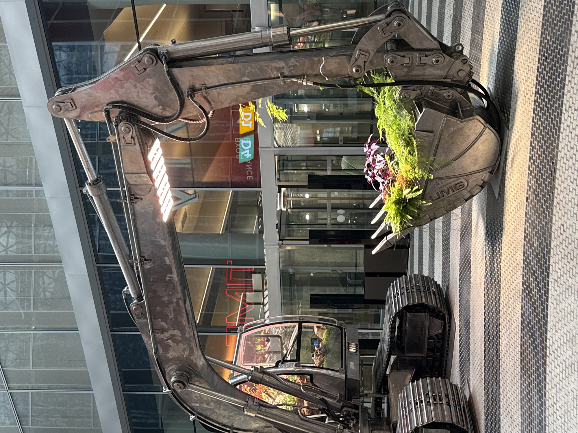 An oversized excavator sculpture, repurposed as a planter, is displayed on a patterned tiled floor in front of a modern glass building. The excavator is made of weathered metal and its bucket is filled with lush green ferns and purple foliage. A grow light is attached to the excavator's arm, illuminating the plants. The building's facade features large glass windows with geometric patterns and reflective surfaces. In the background, revolving glass doors are visible, along with signage in neon and white lettering. The floor is a mosaic of gray and white rectangular tiles, with a distinct striped pattern. The lighting suggests it is daytime, possibly indoors or in a covered outdoor space, given the proximity to the building.