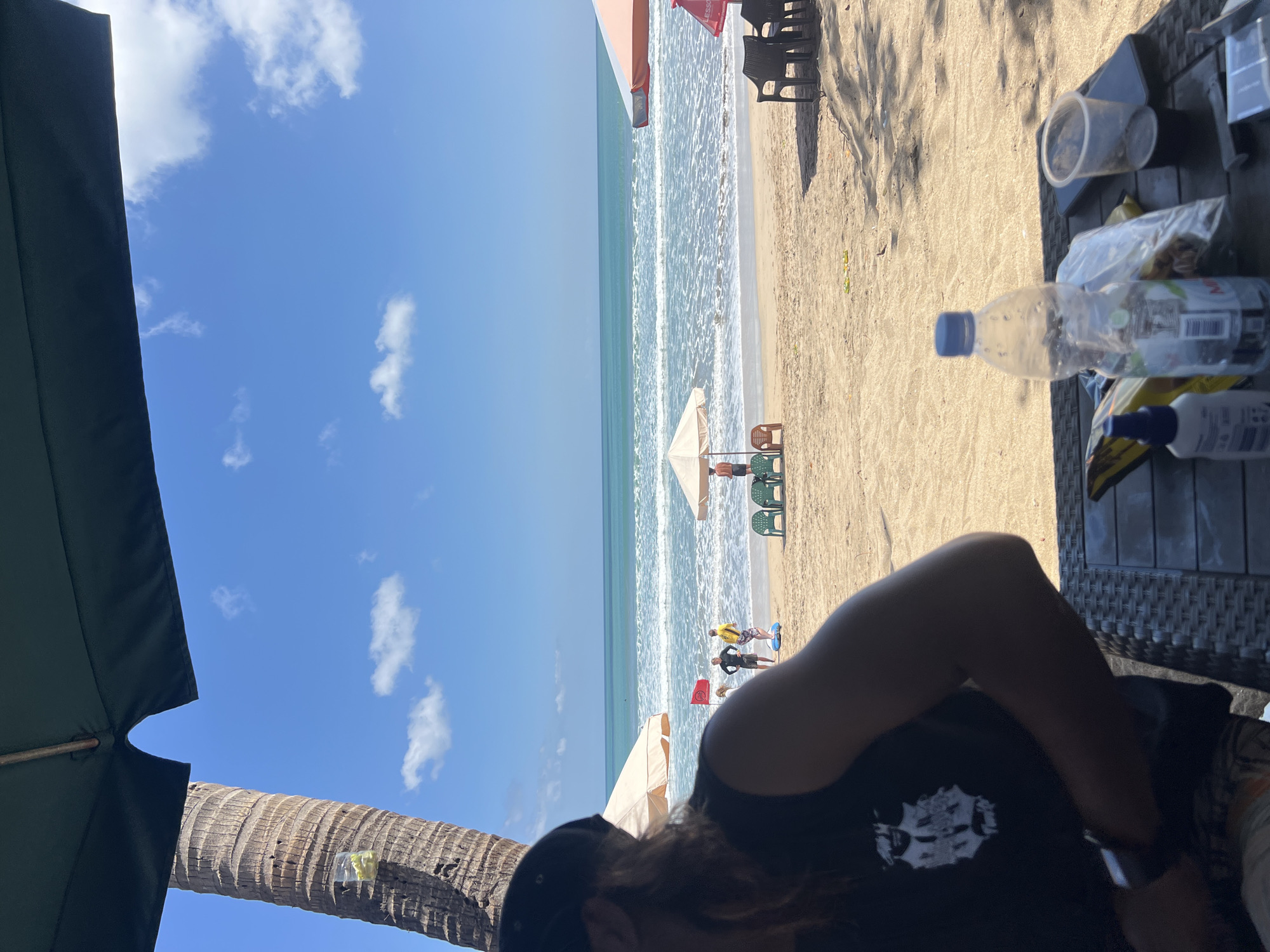 The image depicts a beach scene in Kuta, Indonesia, as viewed from under a large, dark green umbrella. A palm tree trunk is visible to the left, with a plastic cup containing yellow liquid attached to it. The beach is sandy, with the ocean and waves in the background under a bright blue sky with scattered white clouds. Several beach umbrellas, some with chairs underneath, are set up on the sand. A red flag is planted near the water. Three individuals are visible standing on the wet sand near the shoreline, and one person is standing under a separate umbrella further down the beach. In the foreground, a person wearing a black t-shirt and a black baseball cap is partially visible, sitting at a woven table. On the table are a plastic water bottle, a clear plastic cup with a dark liquid, a smartphone, and some plastic-wrapped items. The overall scene conveys a relaxed, sunny day at the beach.