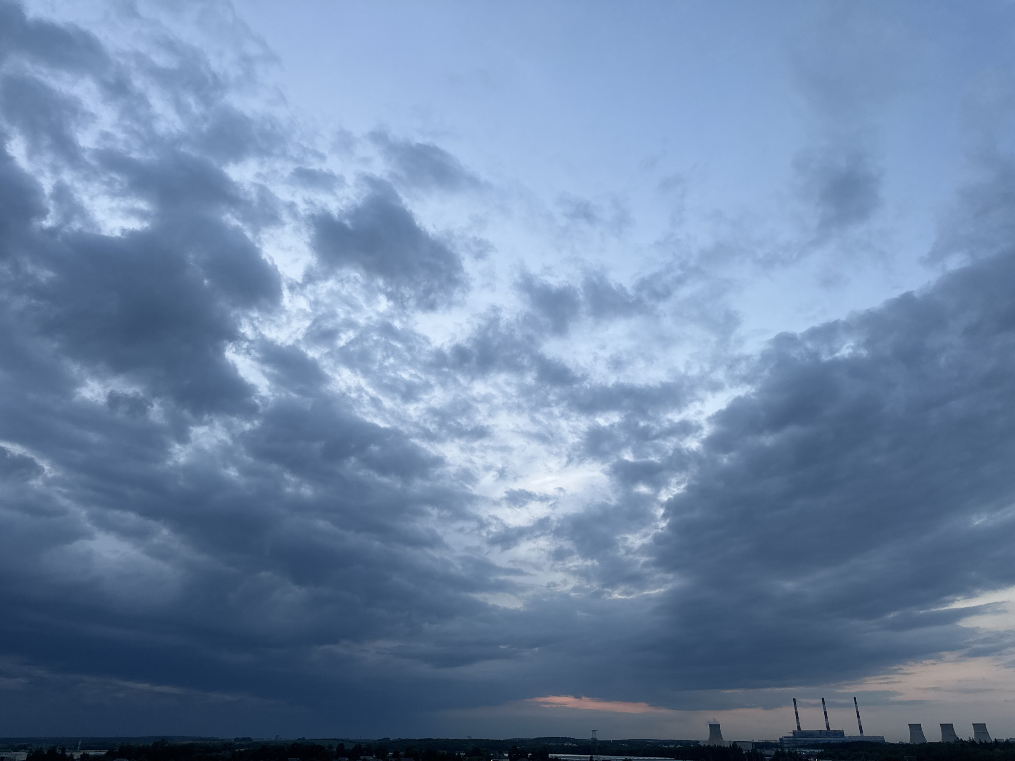The image captures a panoramic view of the sky above Шчомысліцкі сельскі Савет, Belarus. The sky is dominated by a dramatic formation of dark, heavy clouds, suggesting impending rain or a storm. The clouds are varied in texture and density, with some appearing almost black and others a deep gray. Patches of lighter sky peek through, particularly in the upper portions of the image, hinting at a break in the clouds or the faint glow of twilight.
Below the imposing cloudscape, a distant horizon line reveals a landscape dotted with industrial structures. Prominently visible are several tall, cylindrical cooling towers, characteristic of a power plant or industrial facility. These towers stand starkly against the muted sky. Interspersed with the cooling towers are other buildings and structures, suggesting a developed, likely industrial, area within the rural setting. The lower portion of the image is dark, obscuring finer details of the ground level. There are no visible people or active events depicted in the scene. The overall impression is one of a somber, overcast day, with a hint of industrial presence on the horizon.