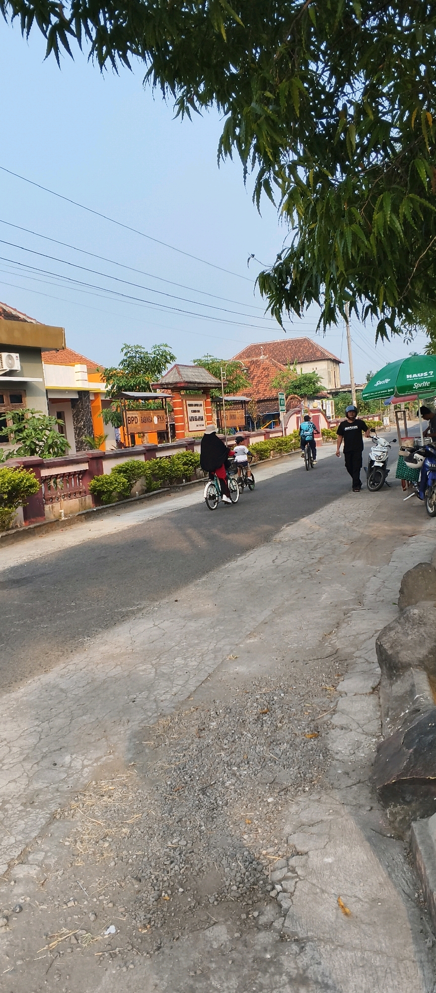 This image depicts a street scene in Japanan, Indonesia. In the foreground, a cracked asphalt road is visible, with a rough concrete embankment on the right side. Several people are present: two individuals on bicycles are in the middle ground, with one person in a black hijab and the other in a white shirt and shorts. Further down the road, a man in a black t-shirt and dark pants walks towards the right, and another person in a blue shirt is on a bicycle. In the background, several buildings with orange roofs are visible, along with some greenery. A green Sprite umbrella stands on the right side of the road, near a food cart. Two motorcycles are parked on the right side of the road. The sky is clear and blue, with tree branches hanging into the frame from the top left and top right.