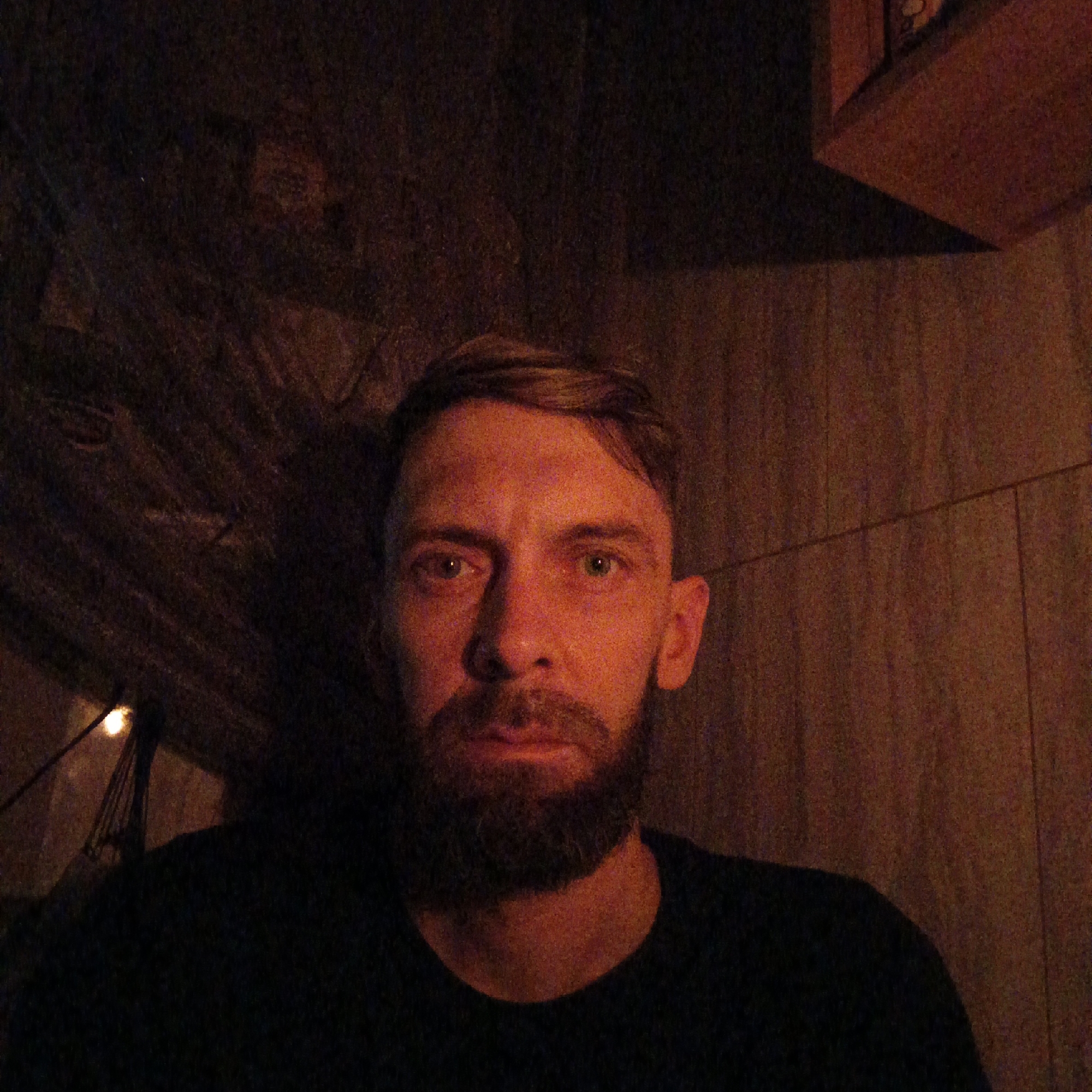 A man with short, light-brown hair and a beard is facing the camera. The lighting is dim, casting a reddish-brown hue on his face. He's wearing a dark shirt. Behind him is a dark, textured wall, and a corner of a shelf is visible in the upper right. A faint light fixture is also visible to the left. The location is specified to be in Марковский, Russia.