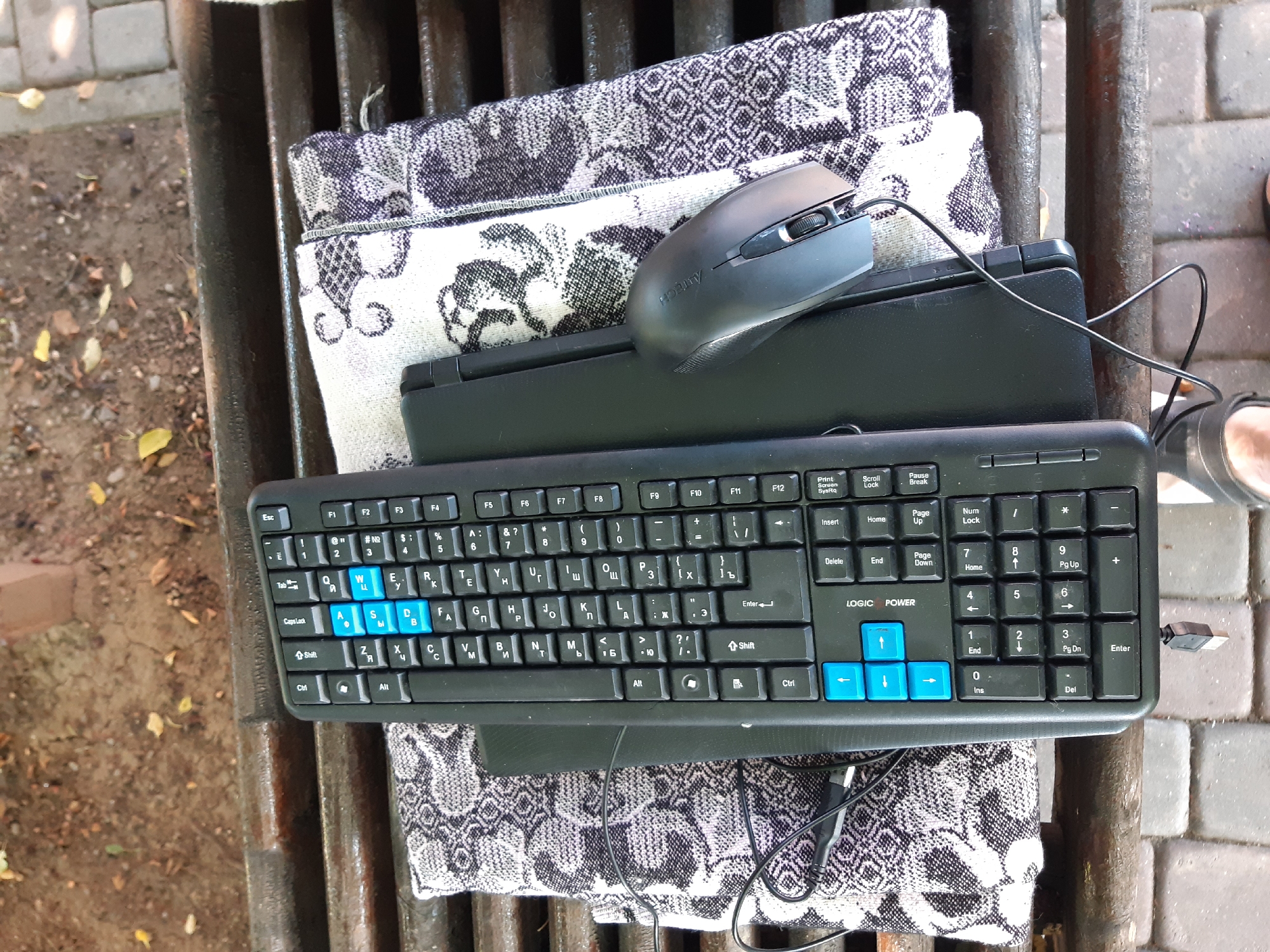 This media file displays a top-down view of a black keyboard and a grey computer mouse resting on a patterned cloth, which is itself on a slatted wooden surface. The setting appears to be outdoors, with hints of cobblestone and soil visible in the background, suggesting a park bench or outdoor seating area. The cloth has a repeating abstract pattern in black and white. The keyboard is a standard QWERTY layout with some Cyrillic characters present, indicating it might be a bilingual keyboard. The keys "W", "A", "S", and "D" are highlighted in blue, often used in gaming. The mouse is positioned on top of a flat black object, possibly a laptop or a laptop case, which is partially covered by the patterned cloth. A black wire snakes from the mouse across the keyboard. The overall lighting suggests it's daytime, likely outdoors. There are no people clearly visible in the frame, though a sliver of a person's foot in a sandal is present on the right edge of the image, adding to the outdoor context. The text "LOGIC POWER" is visible on the keyboard.