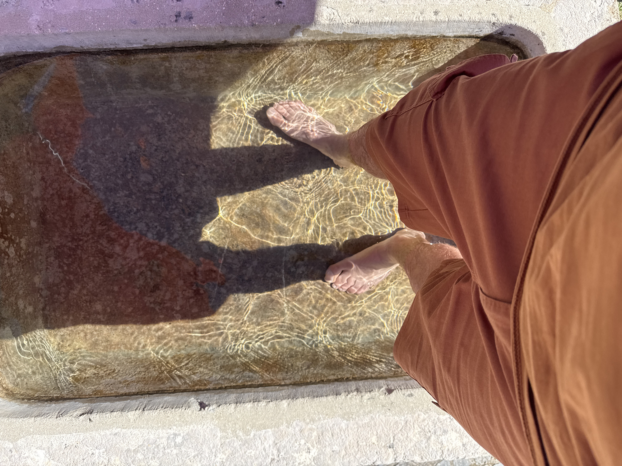 An overhead, close-up shot shows a person's legs and feet submerged in shallow, clear water in what appears to be an outdoor foot spa or water trough. The water is illuminated by sunlight, creating rippling patterns and reflections on the submerged surfaces. The person is wearing reddish-brown shorts. The sides of the spa are constructed of rough, light-colored concrete. A shadow of the person's upper body and head falls across the water. The location of Petit-Val, Switzerland, is not discernible from the image.