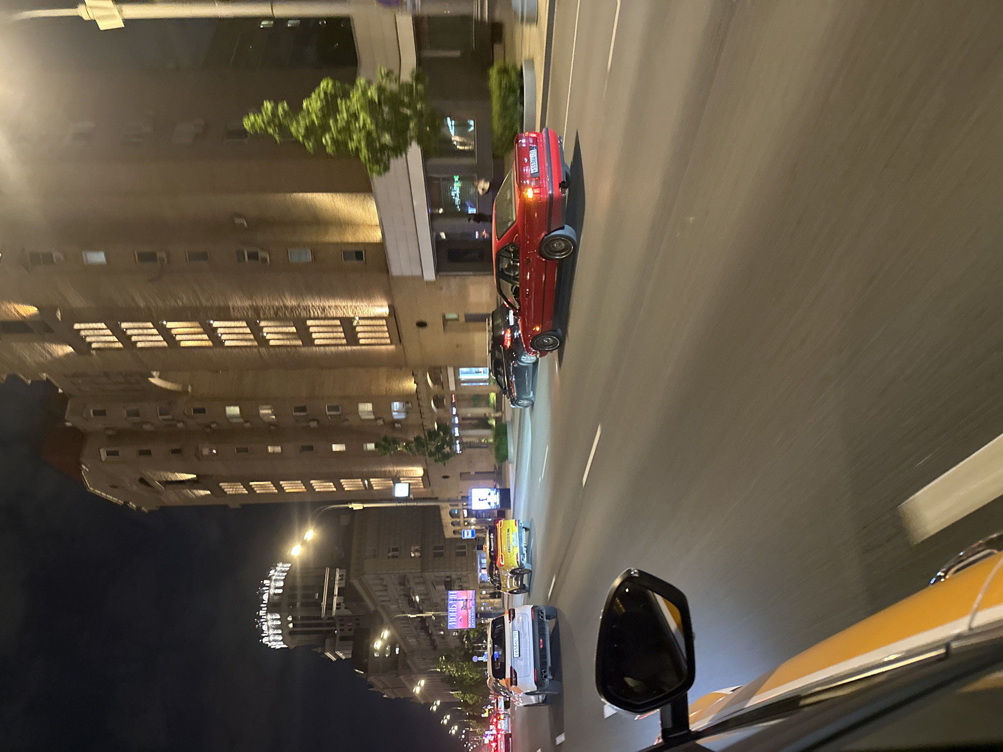The image shows a street in Moscow, Russia, at night. The buildings are illuminated. Cars, including a red vintage car and a yellow taxi, are moving along the road. Street lights are on. The sky is dark. There are billboards along the side of the road.