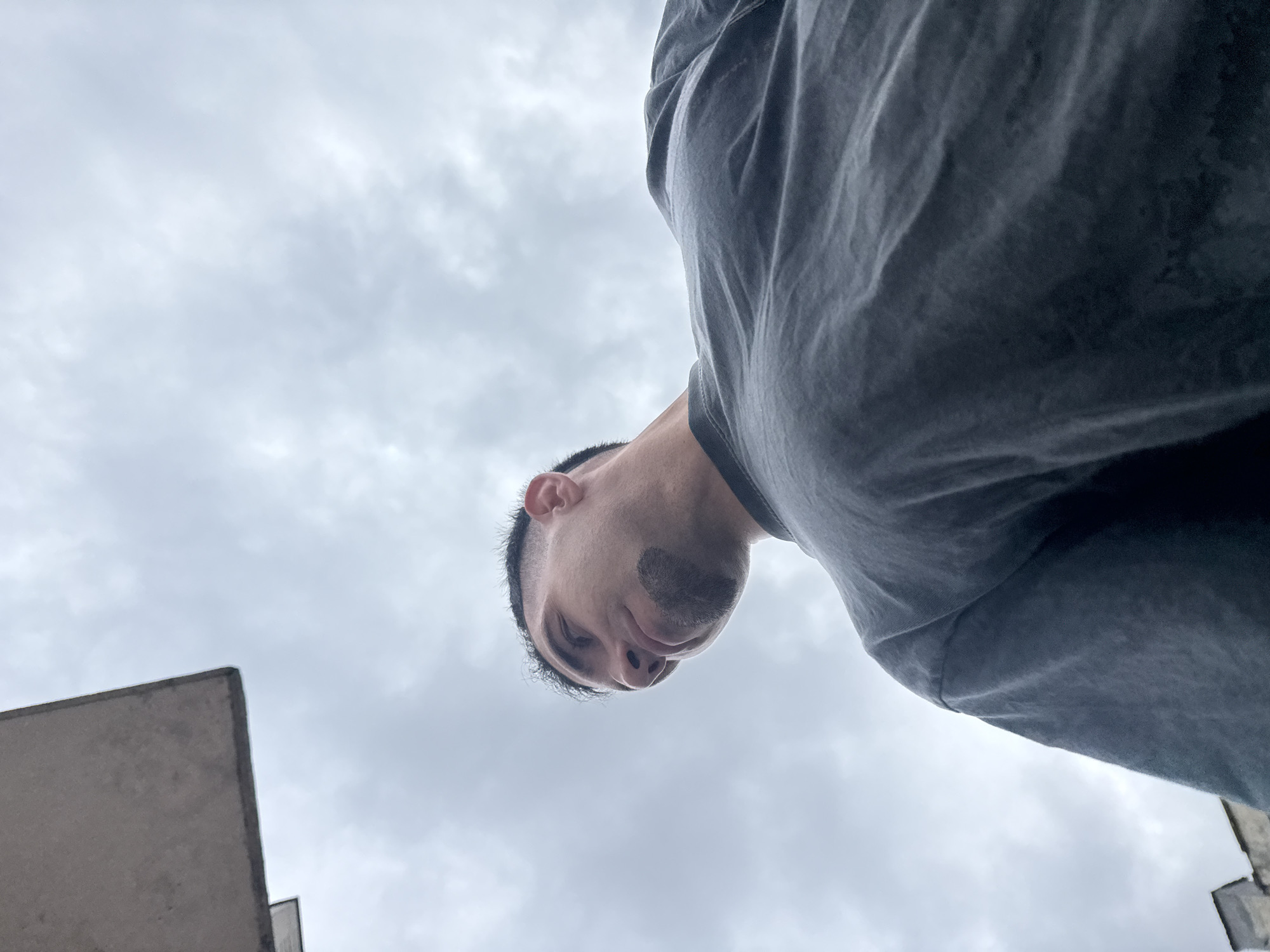 The image shows a low-angle view looking up at a person. The person has dark hair, and facial hair, and is wearing a gray shirt. The background consists of a cloudy sky and a building with concrete elements visible in the lower left corner. The location cannot be identified as Shumen, Bulgaria.