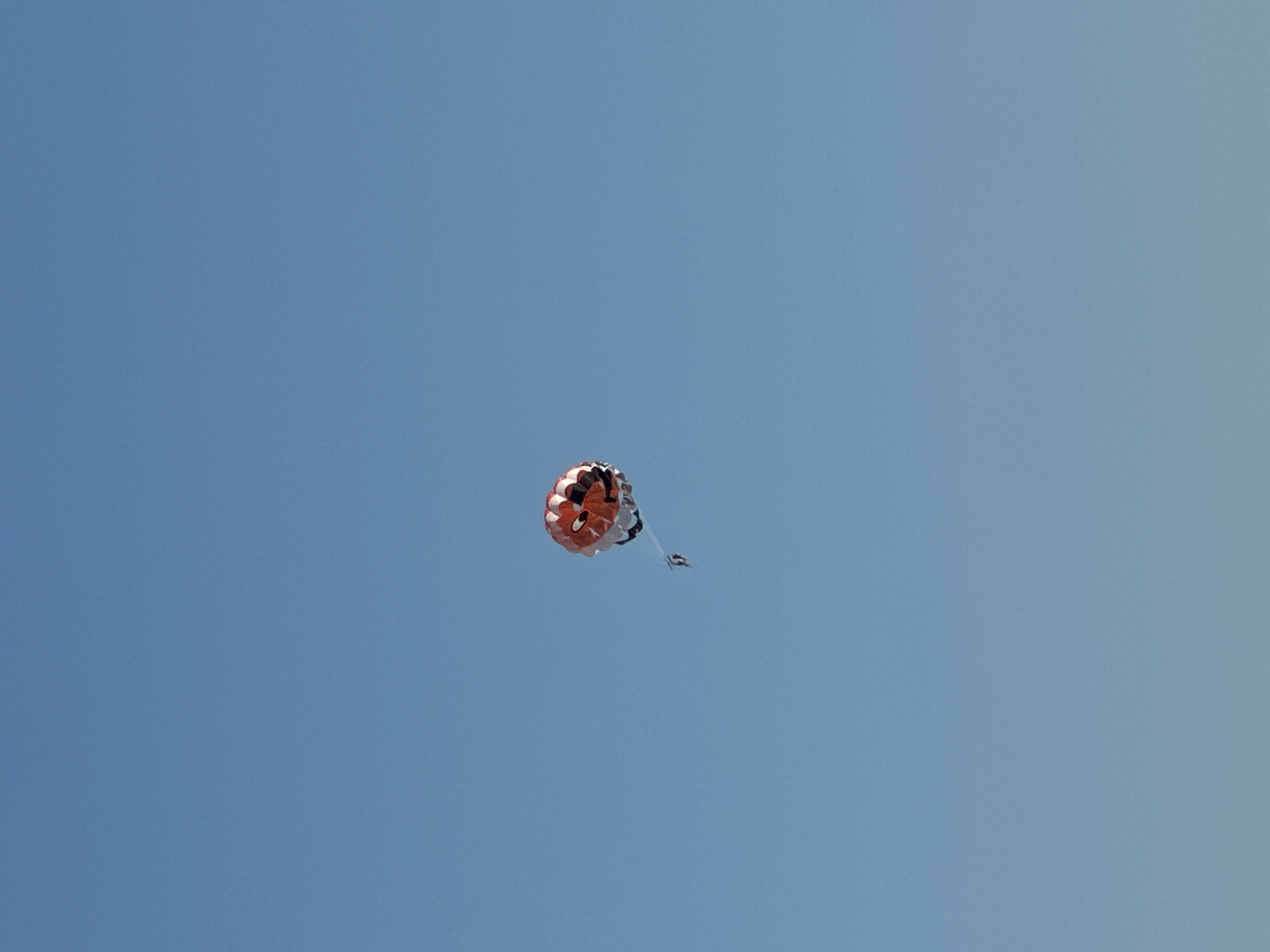 A parasailer is suspended against a clear blue sky, with the parasail shaped like a cartoon character's face, featuring large red eyes and a wide black mouth. The parasail is predominantly white and red. A small figure, the parasailer, is attached by ropes to the parasail. The image captures a moment of aerial activity, likely over water, though the water is not visible in this frame. The scene is devoid of any urban structures or identifiable landmarks that would confirm a specific location within Boreti, Montenegro.