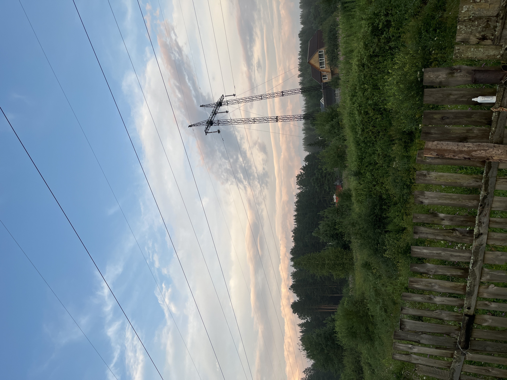 The image depicts a rural landscape under a cloudy sky with a series of high-voltage power lines crisscrossing the frame. In the mid-ground, a tall, lattice-style power pylon stands prominently, leaning slightly. Behind the pylon, a dense forest of dark green trees forms a backdrop. A cluster of houses with wooden facades and visible windows are nestled among the trees. In the foreground, a weathered, wooden fence made of vertical planks runs horizontally across the bottom of the image. The environment appears to be a natural setting, possibly the outskirts of a settlement. This scene is not specific to Nizhny Tagil, Russia, as it lacks identifying landmarks or features that would confirm its location within that city.