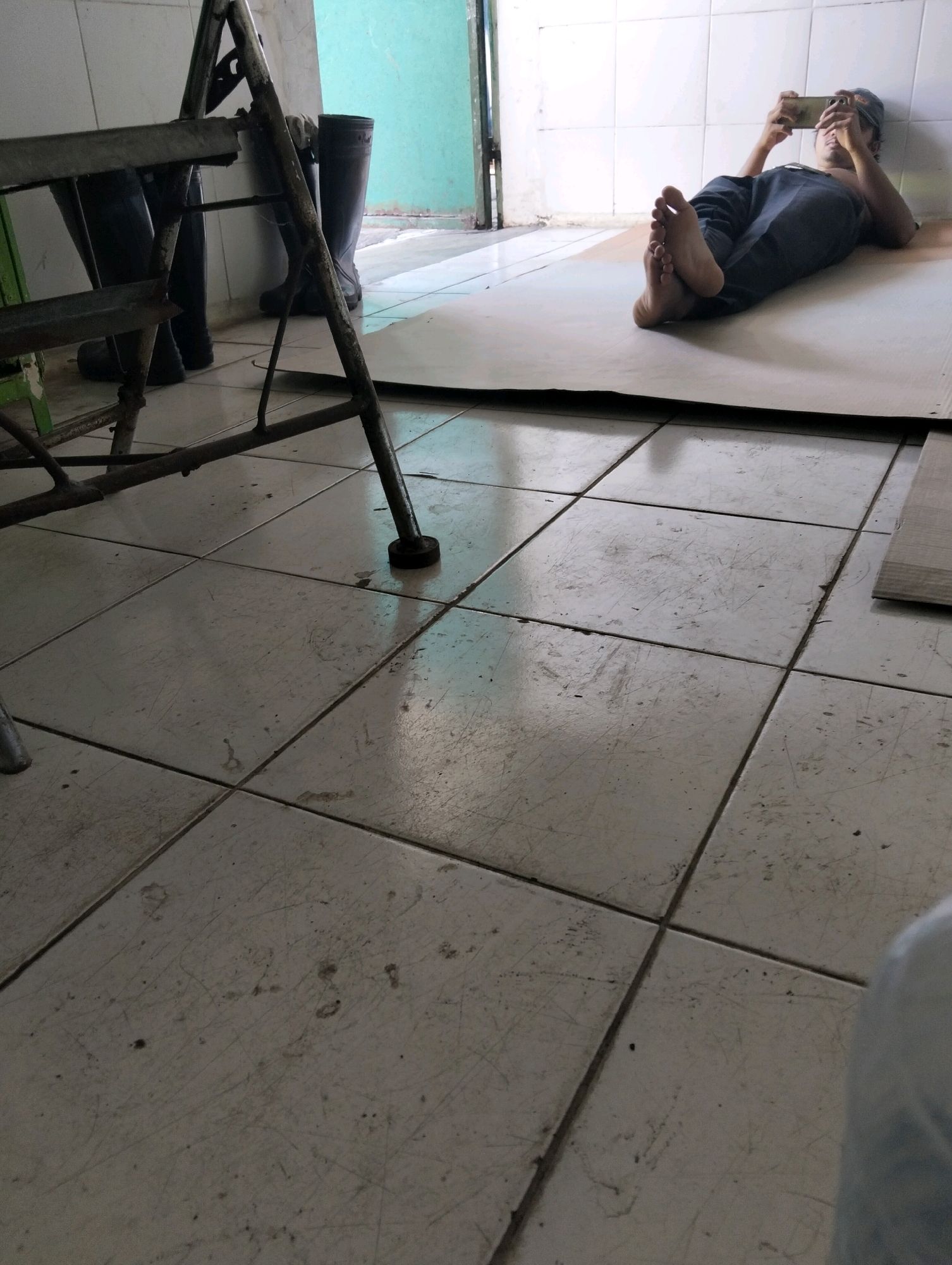 A person, wearing a dark cap and dark pants, is lying on their back on a light-colored sheet of material, leaning against a tiled wall. Their feet are pointed towards the camera. They are holding a smartphone in both hands, looking at the screen. To the left of the person, a metal A-frame ladder is partially visible. Behind the ladder are two dark rubber boots. The floor is tiled in a grid pattern and appears worn. The environment suggests an indoor space, possibly a storage area or a less-maintained room, with elements of a utilitarian setting. The overall scene is candid and captures a moment of rest or leisure.