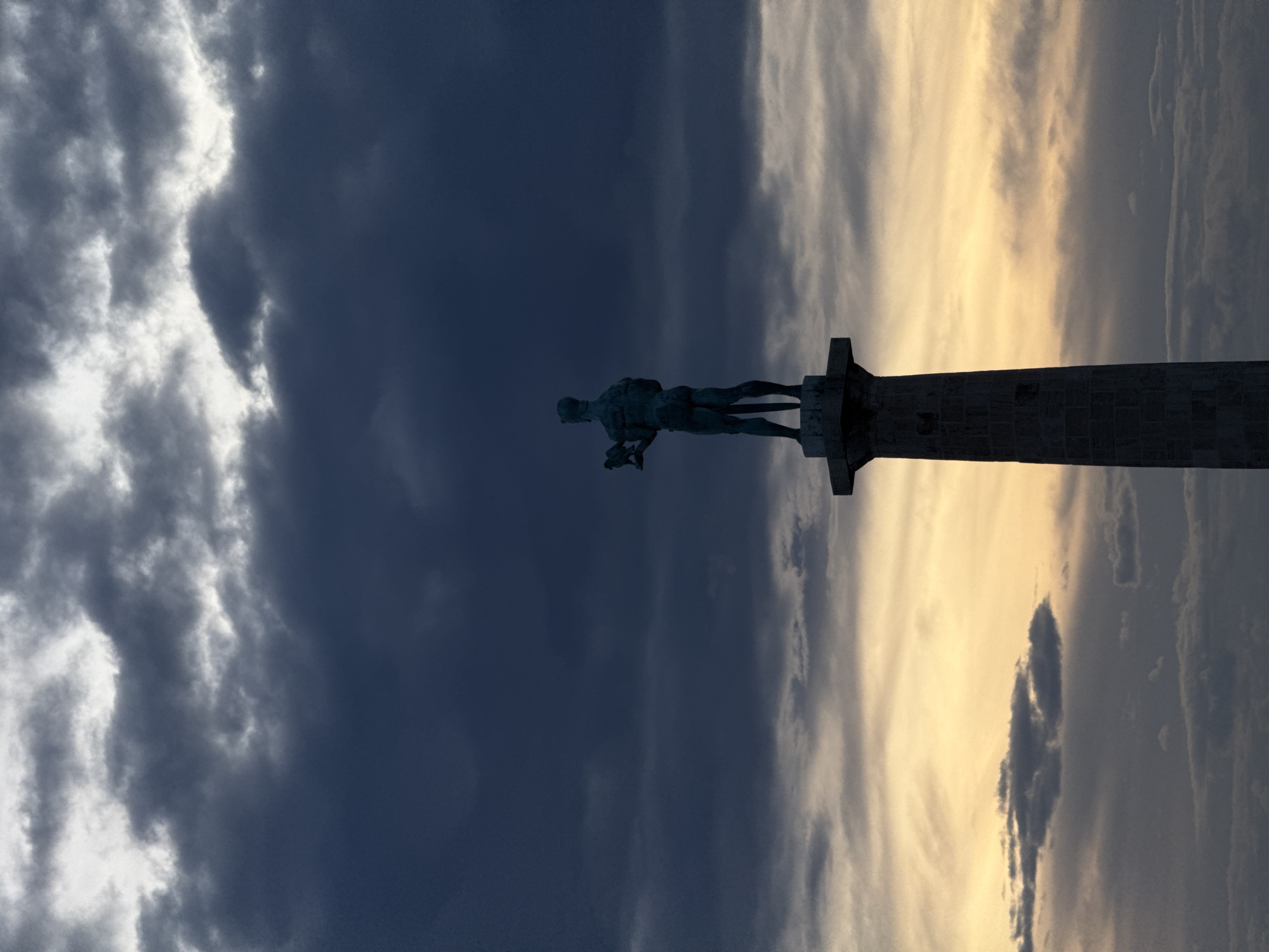 The image shows the Victor monument (Pobednik) atop a pillar in Belgrade, Serbia. The monument is silhouetted against a cloudy sky, with dark, heavy clouds to the left and lighter, sunlit clouds to the right. The statue is male, nude, and holding a sword and a bird (typically interpreted as a falcon). The pillar on which the statue stands is composed of stone blocks. The orientation of the image is rotated.