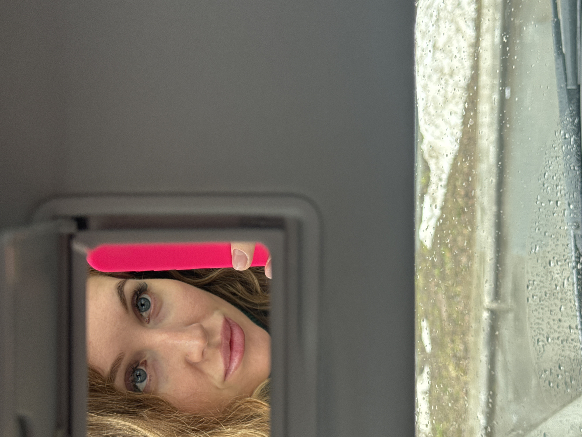 A woman looks at herself in the sun visor mirror of a car. She is holding a bright pink object in her right hand. Raindrops are visible on the car window. The location is Chaikovsky, Russia, based on the request.