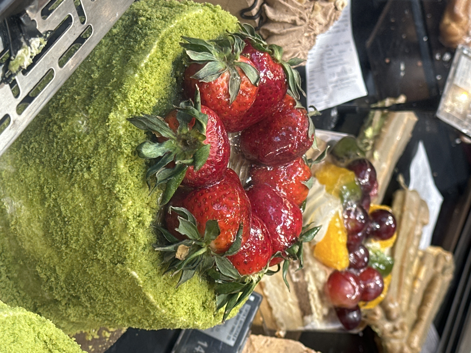 The image shows a close-up view of a dessert display, likely inside a pastry shop or bakery. A vibrant green cake roll covered in what appears to be matcha powder or a similar ingredient is prominently featured, adorned with fresh strawberries on top. Next to it is a dessert with grapes and orange slices, possibly a cake or pastry as well. The desserts are presented behind a glass display case. The image also reveals a metallic, perforated sheet, possibly part of the store's refrigeration or display system. The presence of a price tag suggests the scene is likely within a commercial environment like a shop. Without additional information, the location within the city of Sofia, Bulgaria, cannot be confirmed.