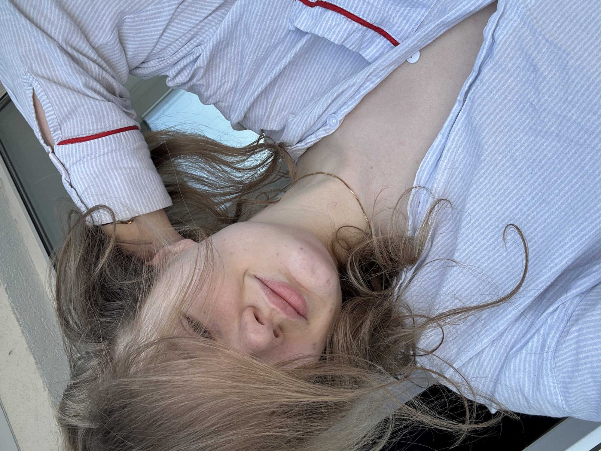 A fair-skinned person with blonde hair is lying down, wearing a light blue and white striped pajama top with red trim. The person is close to the camera, with their face slightly angled. A gold chain necklace is visible. The location cannot be determined from the image.