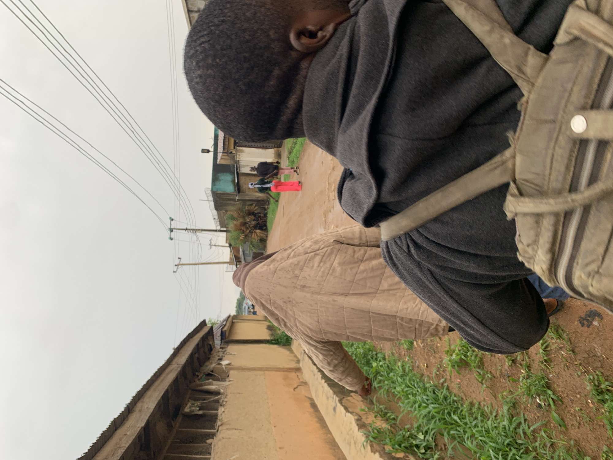 This media file captures a street scene in Rafin Par, Nigeria, under an overcast, grey sky. The image is taken from a slightly elevated perspective, looking down a muddy, unpaved road.
In the immediate foreground, on the right side of the frame, the back of a person's head, right ear, and shoulders are visible. This individual has short dark hair and is wearing a dark grey hooded top. A light brown or khaki backpack strap is visible over their right shoulder.
Ahead of this foreground figure, another person is seen walking away, clad in a light brown quilted jacket. Further down the street, two more individuals are discernable in the distance: one in dark clothing and another wearing bright red trousers or a skirt with a lighter top.
The road itself is a prominent feature, appearing wet and muddy, flanked on the right by a concrete drainage ditch or gutter. Green grass and weeds grow along the edges of the ditch. Utility poles carrying multiple power lines stretch across the expansive, cloudy sky. Simple buildings, some featuring corrugated iron roofs or walls, line both sides of the street. A street light is also visible in the background. The scene suggests a modest, semi-urban or rural environment, likely during the day on a cloudy or recently rainy day. No text is visible in the image.