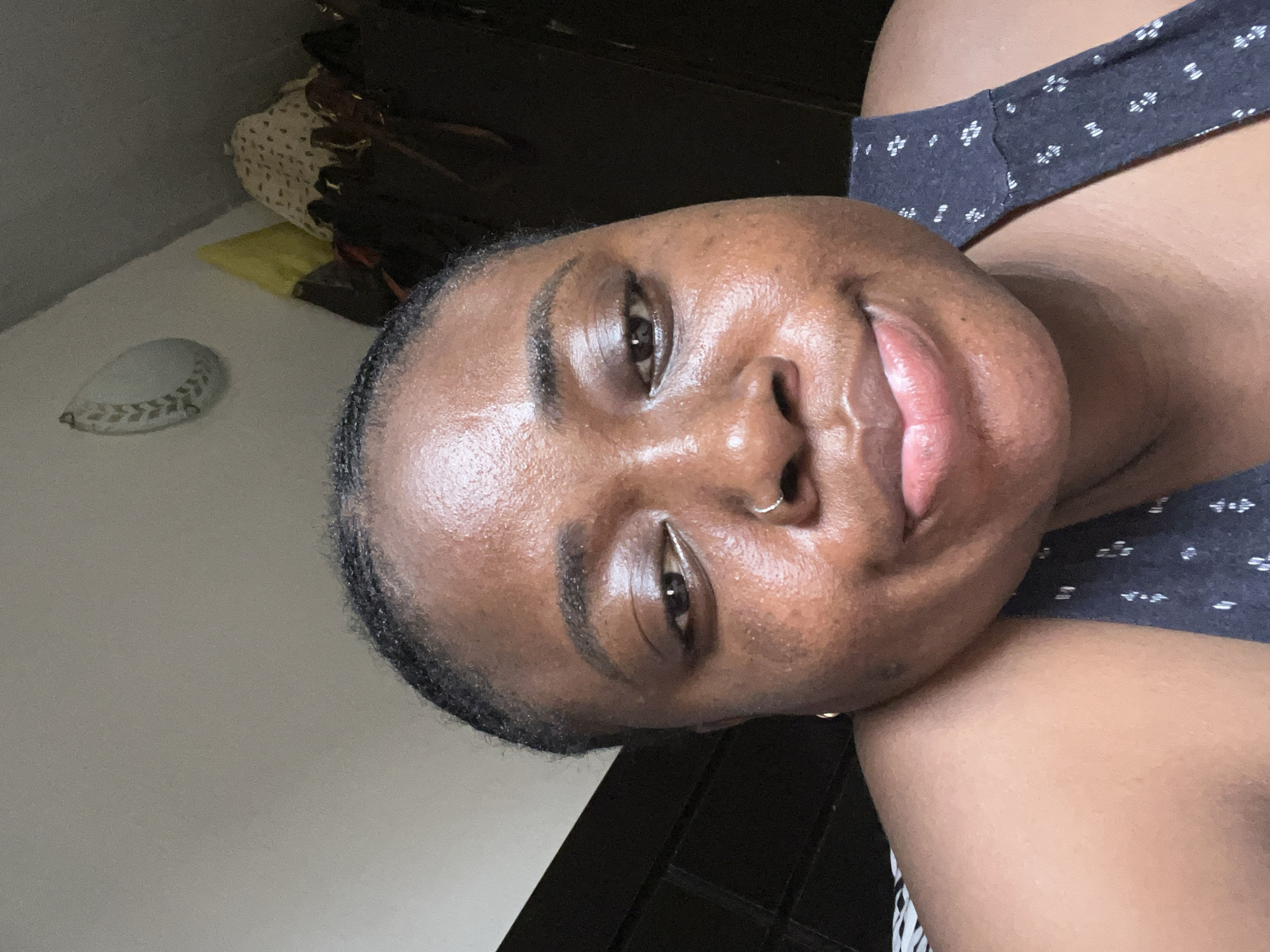 A close-up photograph features a dark-skinned woman with a nose ring, smiling at the camera. She wears a patterned dark gray top. In the background, there's a dark cabinet or closet, a white baseball, and bedding. The location is not discernible from the image. The city of Abuja, Nigeria cannot be confirmed.