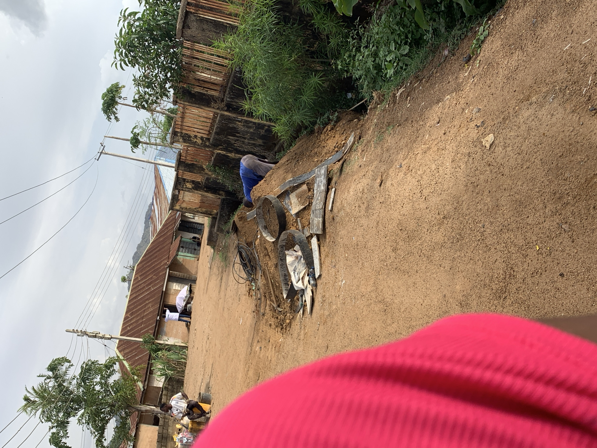 A street scene in Rafin Par, Nigeria shows a sandy road with a person in blue clothing working on the side of the road near some tires and other debris. To the left, there are buildings with corrugated metal roofs, and people can be seen standing near one of them. Power lines run along the street. Vegetation is visible along the roadside and on top of a wall. The lower portion of the image is obscured by a bright pink object, likely clothing.