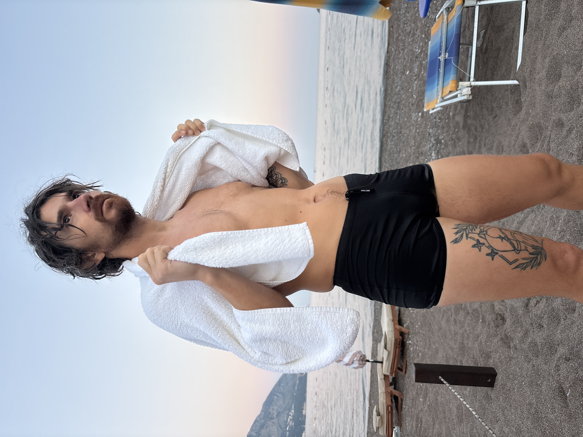 A man stands on a pebble beach in Boreti, Montenegro, wearing black swim trunks and a white towel draped over his shoulders. His hair is dark and slightly disheveled, and he has facial hair. He is looking away from the camera towards the sea. The water is calm, and there are a few beach chairs and umbrellas in the background, along with a gentle slope of a mountain covered in vegetation. The sky is a soft, hazy blue, indicating either early morning or late evening. There is a tattoo visible on his right thigh, depicting a circular design with leaves and stars. Another tattoo is partially visible on his left arm.