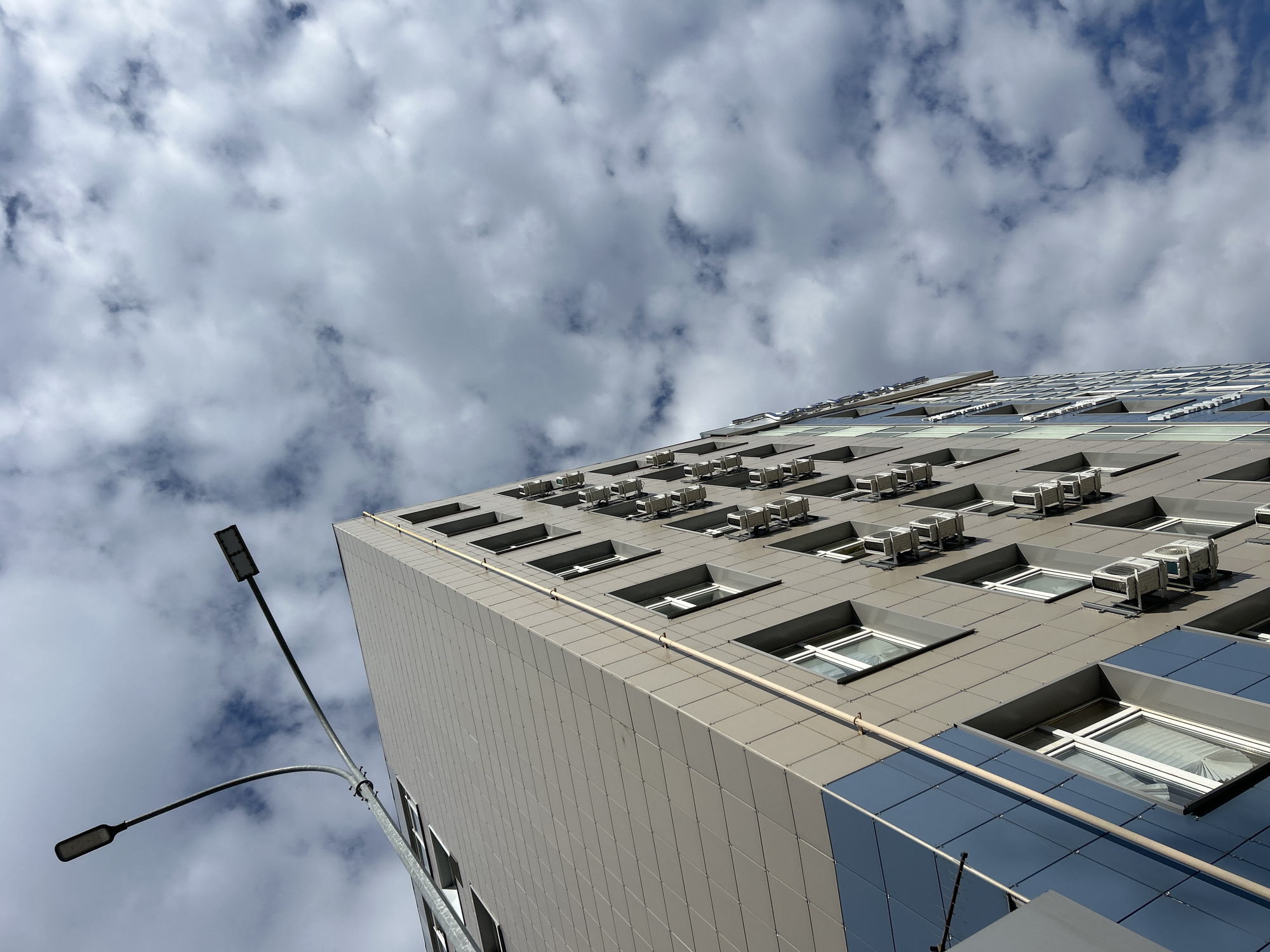An upward-angled view captures a modern multi-story building against a partly cloudy sky in Chaikovsky, Russia. The building's facade is clad in rectangular grey and beige panels, with sections of dark blue reflective glass on its lower right side. Numerous air conditioning units are visible, mounted below recessed, rectangular windows across several floors. A light-colored pipe runs horizontally along one of the building's paneled sections. To the left, a tall street light pole with two modern, rectangular LED-style lamps extends into the frame, with one lamp pointing higher and the other angled downwards. The sky is dominated by a mix of white and grey clouds, with glimpses of clear blue. The bright, natural lighting indicates it is daytime. There is no discernible activity or visible text in the image. The scene portrays a contemporary urban environment.