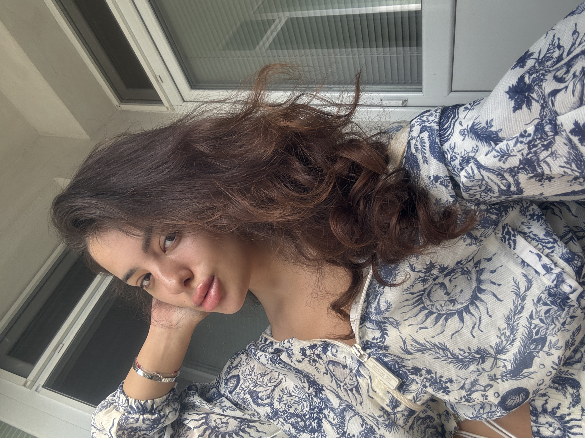 A young woman with long, dark brown, wavy hair is shown in a selfie, with her left arm bent and hand behind her head. She is wearing a white top with a blue toile pattern, featuring motifs of suns, animals, and foliage. A silver bracelet adorns her left wrist. Her gaze is directed at the camera, and her lips are slightly parted, giving the impression of a kiss or a pout.
The background suggests an urban environment, possibly a balcony or an exterior wall of a building. A closed window with horizontal blinds is visible behind her right shoulder. The architecture appears to be typical of Plovdiv, Bulgaria, characterized by its light-colored facades and modern window frames. The overall lighting indicates it is daytime, with sunlight casting subtle shadows and highlighting the woman's features.