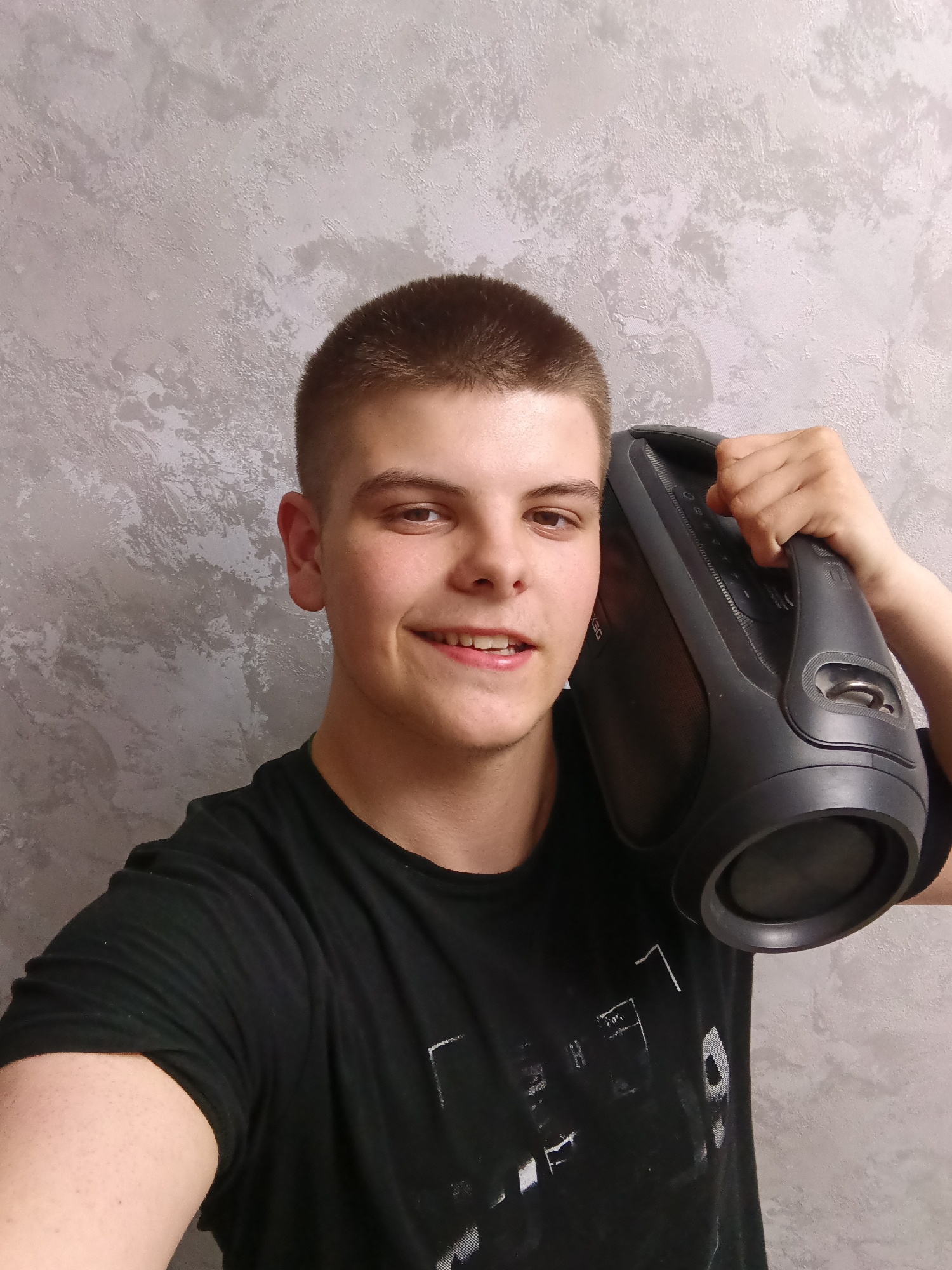 A young man is holding a black speaker on his right shoulder. He is smiling and wearing a black t-shirt. The background is a gray textured wall. The location is Unknown, Unknown.