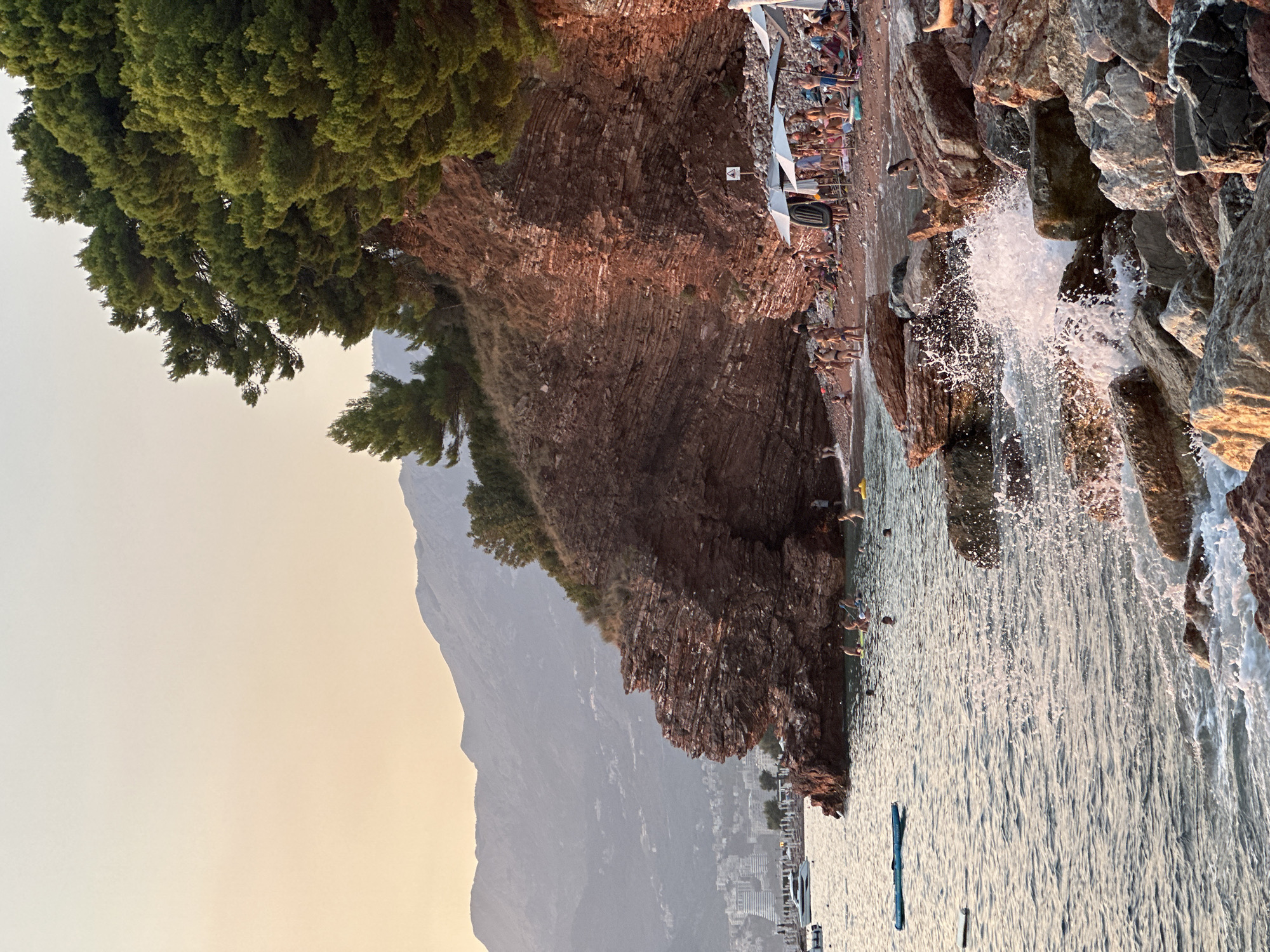 A sunset view of a rocky beach in Sveti Stefan, Montenegro, is captured. The foreground shows waves crashing against the rocks, creating white spray. Further back, a beach is populated with numerous people sunbathing, swimming, and relaxing under umbrellas. Some individuals are in the water, while others are seated or standing on the sand. Behind the beach, a large, layered rock formation rises, topped with lush green pine trees. In the distance, a city with buildings and mountains can be seen, bathed in the soft light of the setting sun. A few boats are visible in the calm water. The overall scene depicts a serene and active coastal resort during the golden hour.