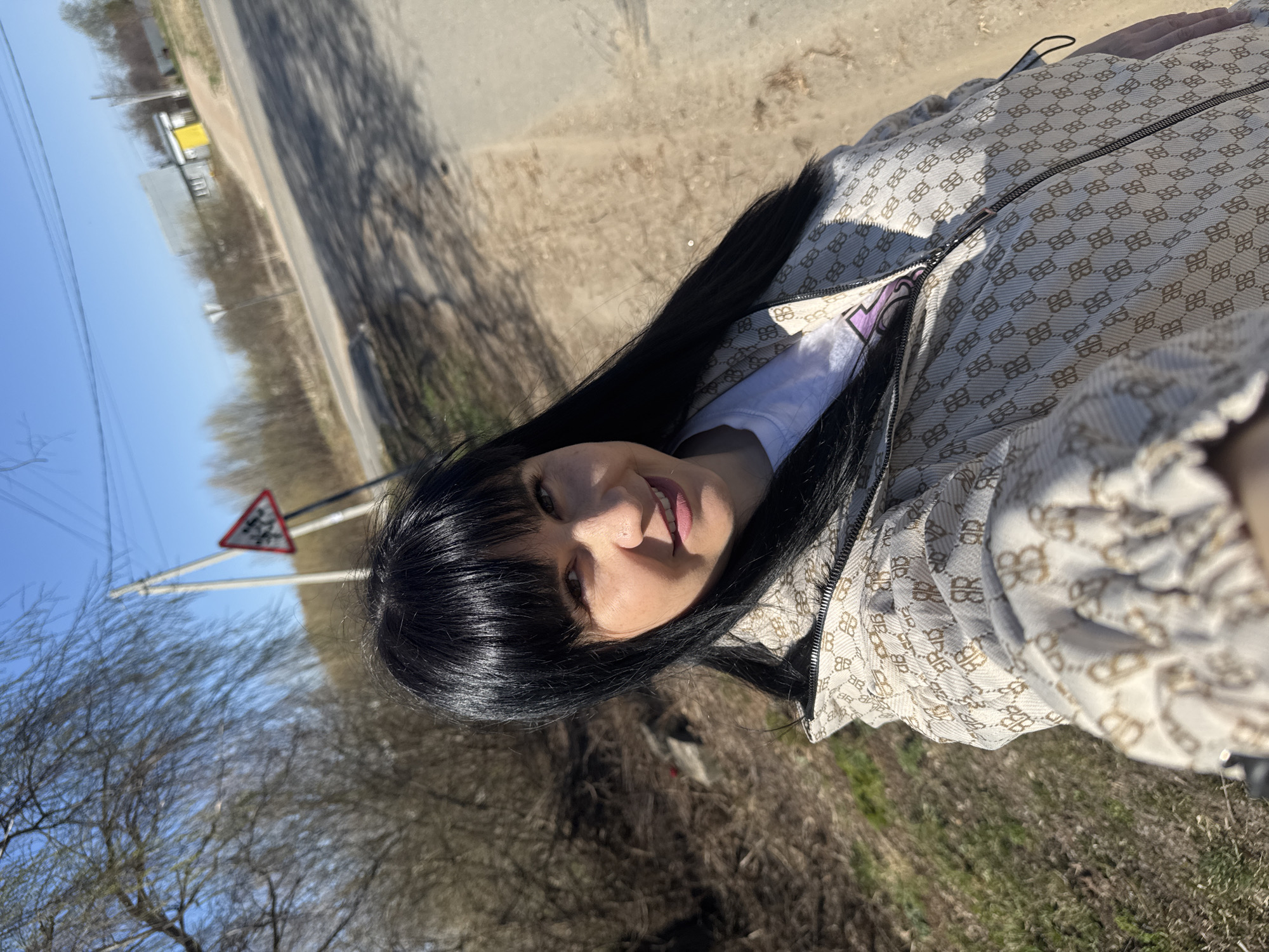 The image shows a woman with long, black hair and bangs, wearing a patterned jacket and a white t-shirt. She is outdoors, near a road, as evidenced by a road sign in the background. The environment consists of bare trees and blue sky. The location might be near a rural road on the outskirts of Zav'yalovo, Russia, as suggested by the open landscape and infrastructure.