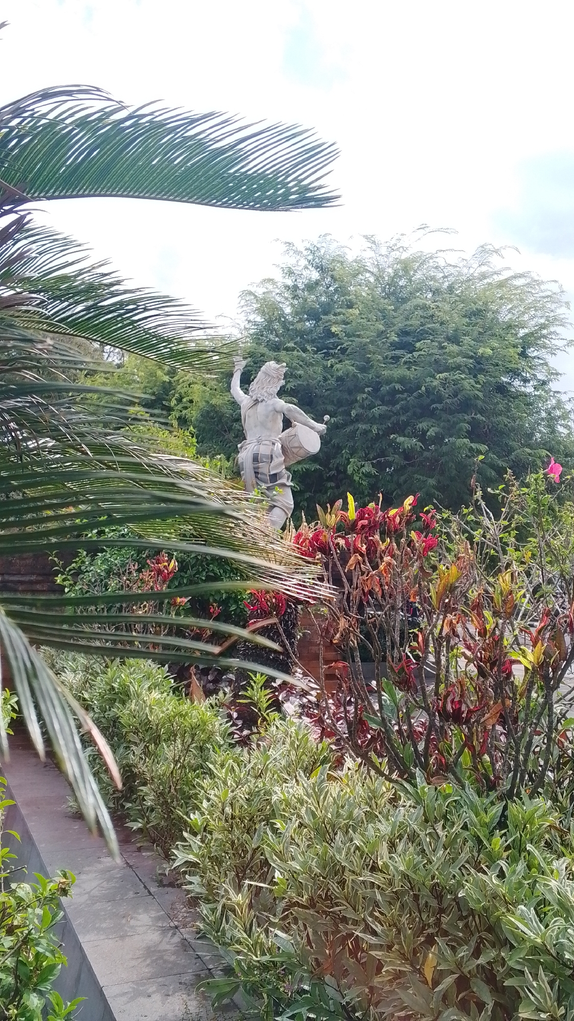 A statue of a person playing a drum stands in a garden. The statue is carved from a light-colored stone and depicts a figure in traditional clothing, possibly playing a Balinese drum. The statue is surrounded by lush green foliage, including palm fronds in the foreground, and a variety of flowering plants with red and green leaves. In the background, a large, dense green tree provides a backdrop. The sky is cloudy. The overall scene suggests an outdoor setting, possibly a park or a temple.
