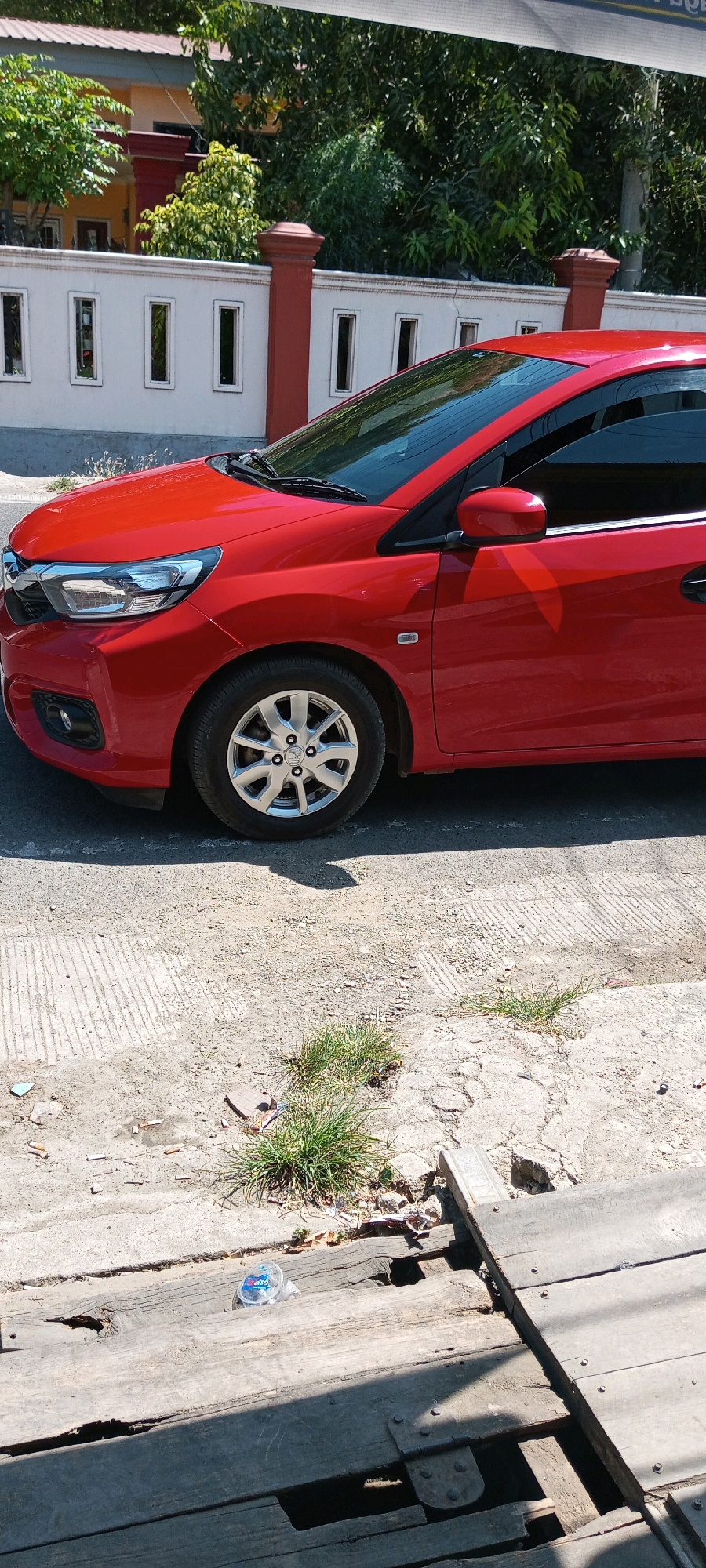 A red car is parked on a cracked concrete surface in front of a white wall with red pillars. Some green grass grows through the cracks in the concrete. A clear plastic bottle is lying on a wooden surface in the foreground. The scene appears to be outdoors, possibly on a street or driveway in Sengkang, Indonesia, as indicated by the presence of a red car, typical residential architecture, and vegetation. The lighting suggests it is daytime.