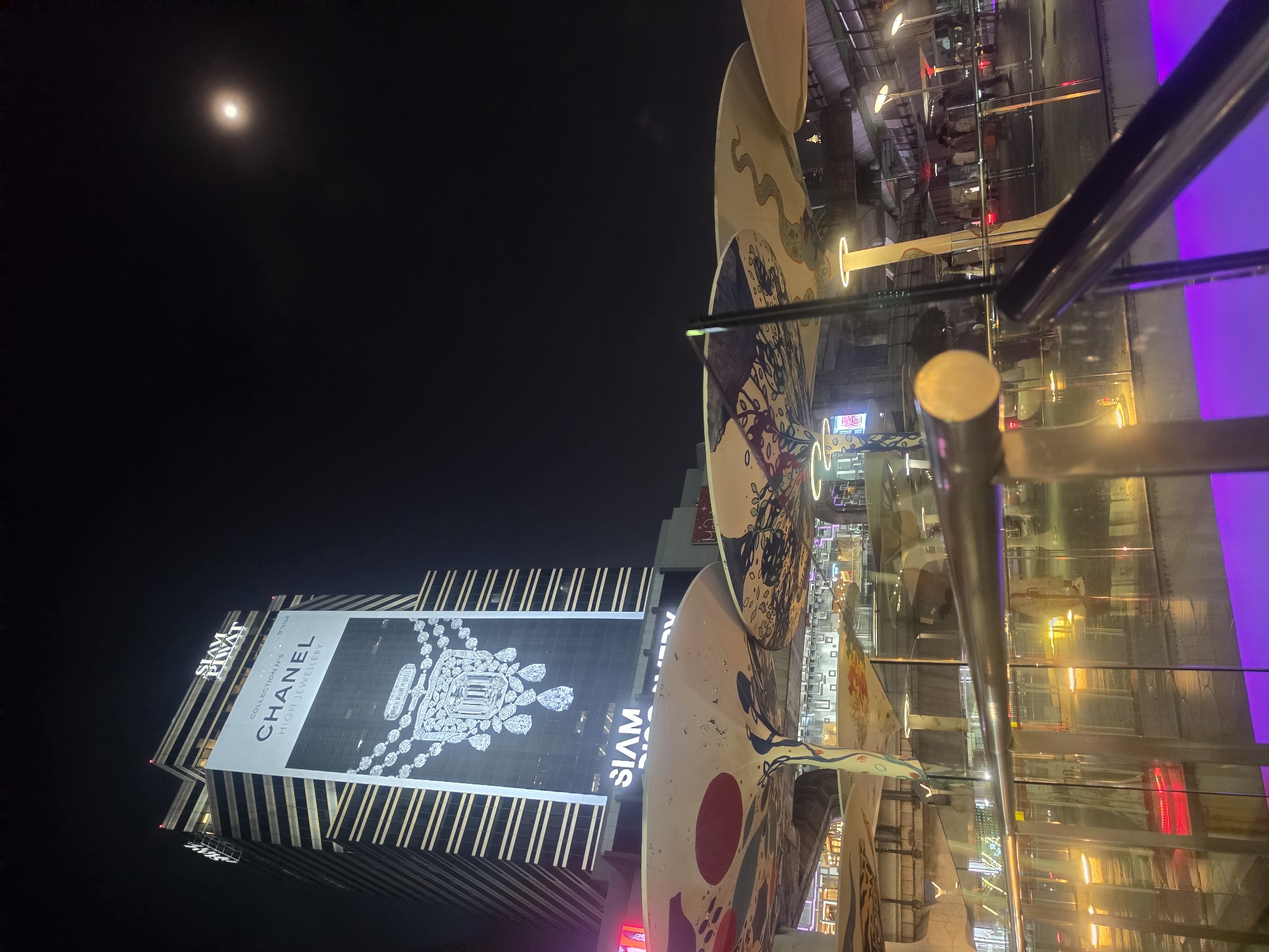 The image presents a nighttime urban landscape, likely in Bangkok, Thailand, viewed from an elevated perspective. A bright, near-full moon is visible in the clear, dark sky in the upper left.
The central subject is a tall, modern skyscraper, dramatically lit against the night. A large banner on its facade prominently features "CHANEL HIGH JEWELRY" with "COLLECTION N°5" and displays an elaborate, sparkling diamond necklace design. Above this, the words "SIAM PIWAT" are visible, and below, partially obscured, is "SIAM". The building's architecture includes a patterned facade with vertical lines.
In the mid-ground, large, curved, disc-like structures, possibly parts of an art installation or architectural decor, are visible. These are adorned with colorful, abstract patterns, including a winding snake motif and circular designs. They reflect the surrounding lights.
The immediate foreground includes metal railings and glass panels, indicating the viewpoint is from a balcony or elevated walkway. Reflections in the glass show blurred figures of people, other buildings, and various artificial lights in hues of yellow, red, and a dominant purple glow on the right side of the frame, hinting at a lively commercial area below. The entire scene is illuminated by artificial lights, creating a vibrant contrast with the dark sky. The visible text "SIAM PIWAT" and "SIAM" are strong location cues for the Siam district in Bangkok.