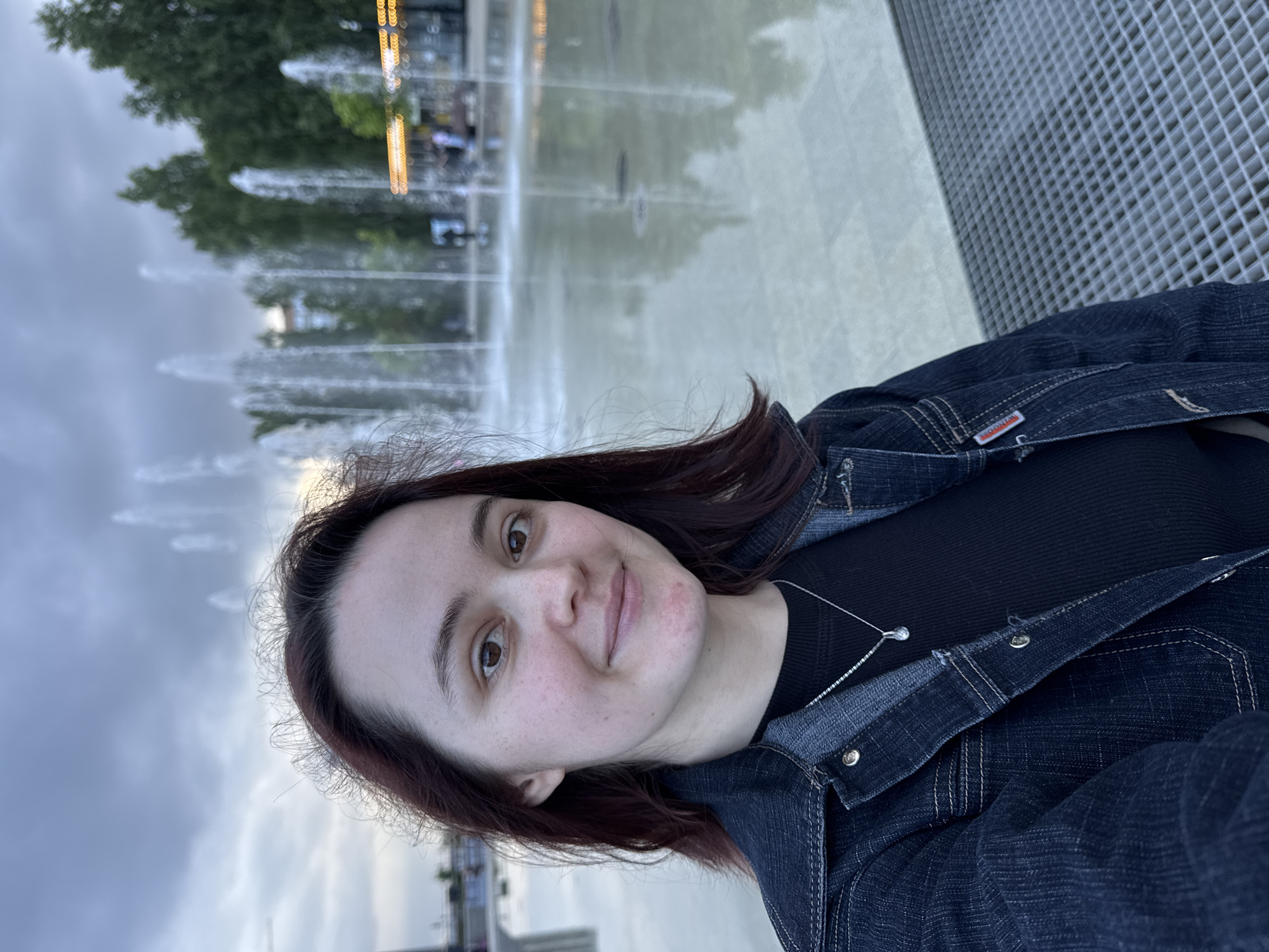 A young woman with dark hair and brown eyes smiles at the camera in a selfie. She is wearing a dark denim jacket over a black top and a silver necklace with a teardrop pendant. In the background, a fountain sprays water, and trees and a building are visible. The scene appears to be a public space with a modern fountain area, possibly in a city park or plaza. The overall lighting suggests it is either early morning or late evening. This location is identified as the "Fountain of Friendship of Peoples" in Izhevsk, Russia.