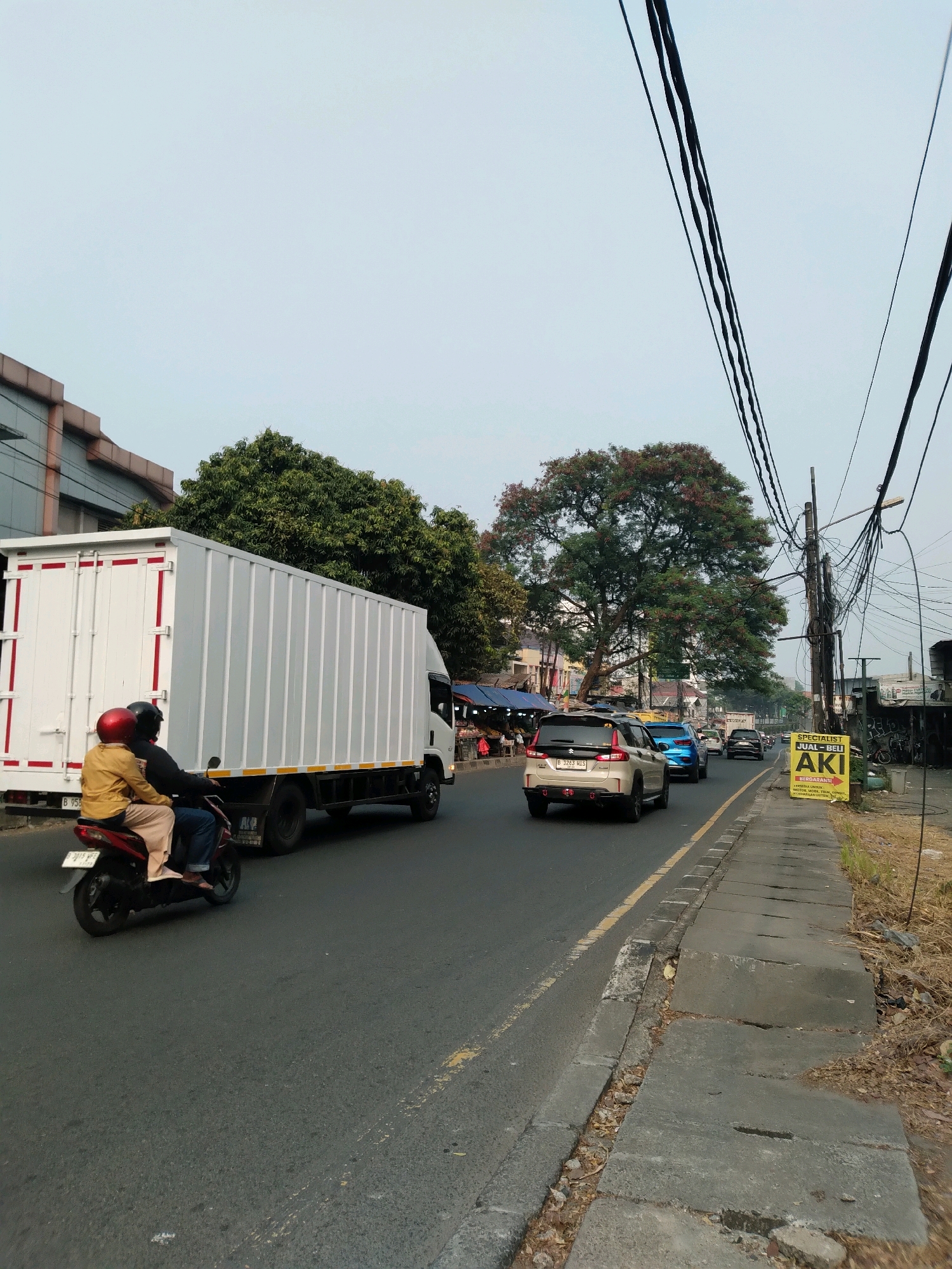 The image depicts a street scene in South Tangerang, Indonesia, under a clear sky. In the foreground, a motorcycle carrying two individuals, a driver wearing a helmet and a passenger in a yellow jacket and light-colored pants, travels down a road. The motorcycle is positioned to the left of a large, white box truck with red and yellow stripes. The truck has a white container and is moving in the same direction as the motorcycle.
Further down the road, several cars are visible, including a silver SUV in the middle lane, followed by a blue sedan and other vehicles. The road is flanked by a sidewalk constructed of concrete slabs, some of which are cracked and uneven. On the right side of the sidewalk, a yellow sign with black text reads "SPECIALIST JUAL-BELI AKI BERGARANSI" and lists types of batteries. Beyond the sidewalk, dry vegetation and debris are present.
The environment includes buildings on the left side of the street, with a modern building featuring a patterned facade. Several trees are scattered throughout the scene, with a large tree bearing reddish flowers dominating the background. Overhead, a complex network of electrical wires and poles is prominently displayed against the sky. In the distance, more buildings and streetlights are visible, indicating an urban or semi-urban setting. The overall scene suggests a typical day with moderate traffic.