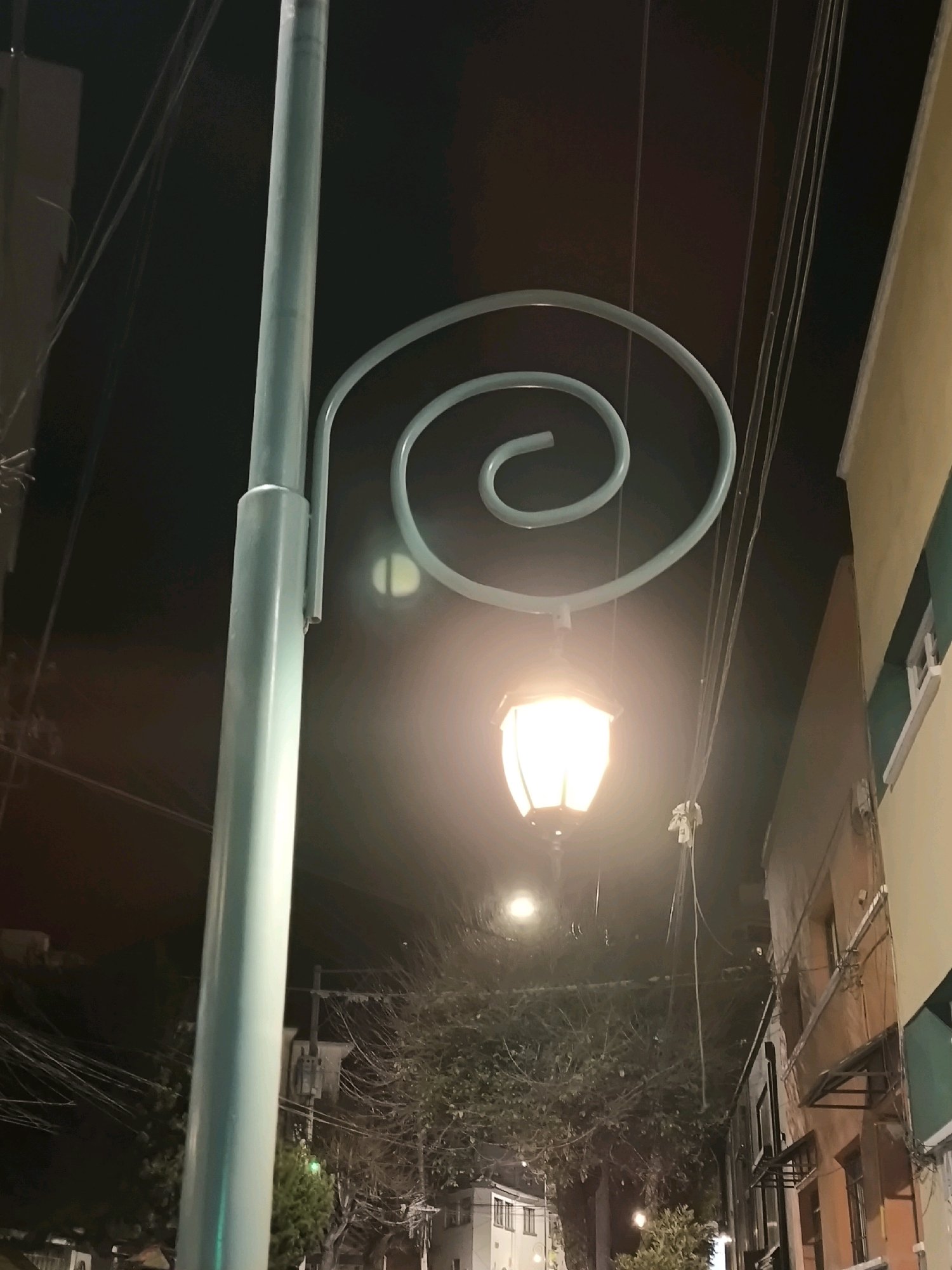 This image shows a streetlamp with a pale blue pole and a spiraling arm, on which a lantern with a warm, glowing light is attached. The streetlamp is located in the city of La Paz, Bolivia. In the background, there are buildings, trees, and overhead electrical wires, typical of a city street at night. The lighting suggests it is nighttime, with the streetlamp being the primary light source in the immediate foreground. The perspective is from a low angle, looking up at the streetlamp.