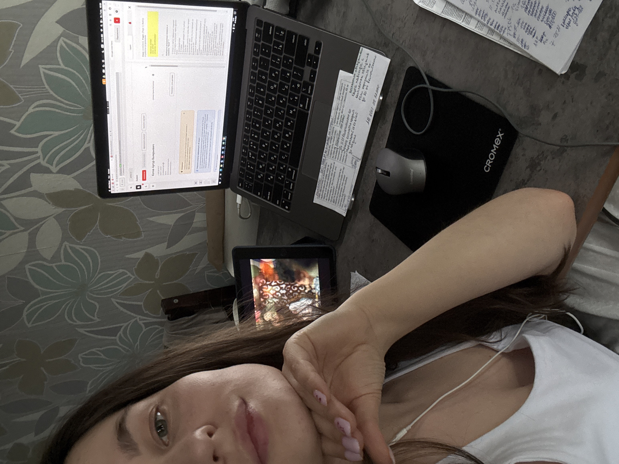 Here is a detailed description of the image:
**General Overview:** The image appears to be a selfie taken by a person with long brown hair, wearing a white top and headphones. The setting seems to be a casual, possibly home-based workspace.
**People:** The primary subject is a person who is likely taking the selfie.
**Subjects and Objects:**
* A laptop is open, displaying a document on the screen.
* There is a mouse on a black mousepad with "CROMEX" printed on it.
* A piece of paper with handwritten notes in Russian is next to the laptop.
* A tablet is visible, displaying a video or image.
* The background includes a floral-patterned surface, likely a wall or wallpaper.
**Actions and Interactions:** The person is taking a selfie, likely while working or relaxing. The devices suggest they might be engaged in work or study.
**Environment:** Based on the Russian text on the paper and the file on the laptop, the image could potentially be in Russia. Without additional elements, it is impossible to pinpoint the location in Chaikovsky, Russia.