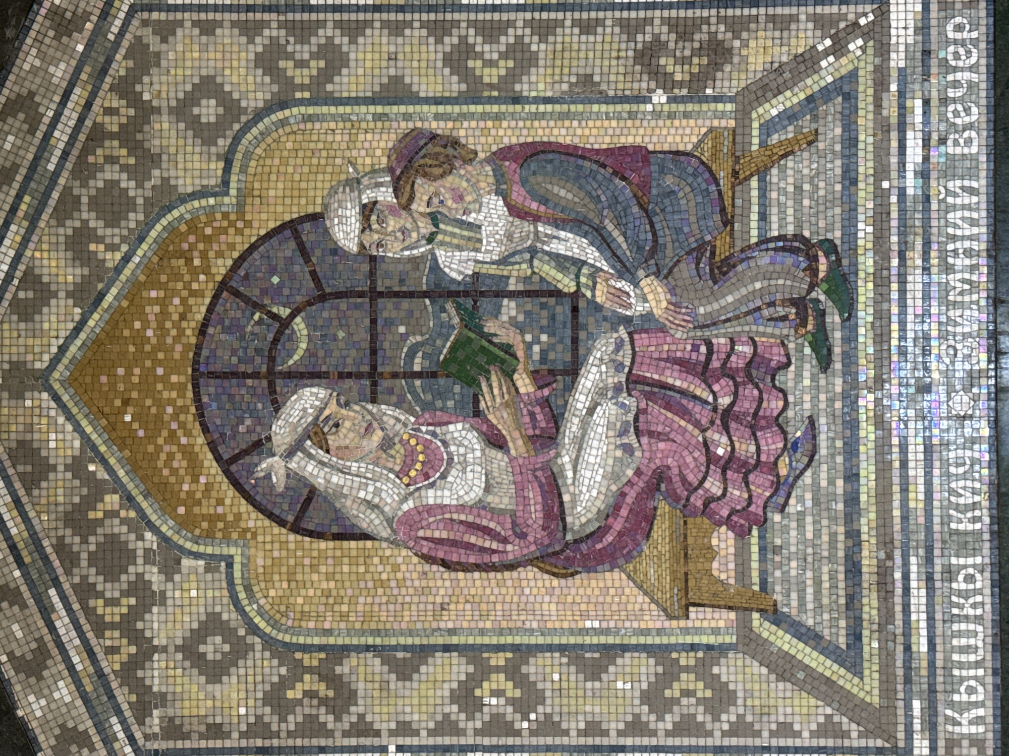 The image is a mosaic panel located in the Kazan Metro station in Kazan, Russia. The mosaic depicts two figures reclining near a window. One figure, possibly female, is lying down and reading a green book. The other figure, appearing male, is resting near the reading figure, with both figures wearing head coverings. The scene is composed of small, square tiles in various shades of color, surrounded by patterned borders. The mosaic also includes the name of the artwork or a relevant text written vertically in Cyrillic.