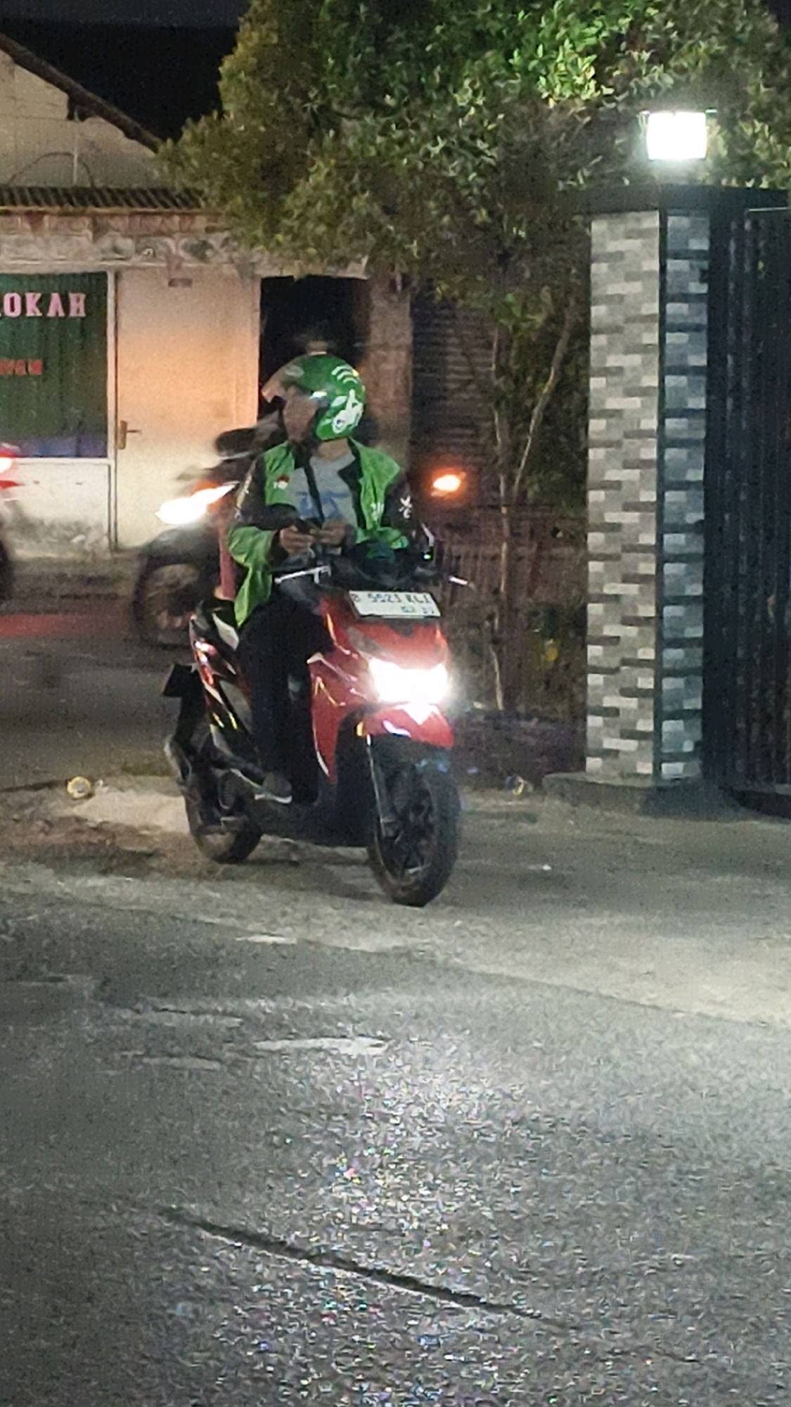 A low-angle, slightly blurry, night time photograph captures a male motorcycle rider in a green jacket with a white logo and a green helmet with a white motorcycle graphic, riding a red scooter. The rider is looking down at a mobile phone he is holding. The scooter has its headlights on, illuminating the wet asphalt road. In the background, there is a building with the word "LOKAH" visible on a green sign. To the right of the building, a tree with green leaves is illuminated by a bright light fixture on a brick pillar, part of a fence. The scene is consistent with a street in Bekasi, Indonesia, likely during or after rainfall.